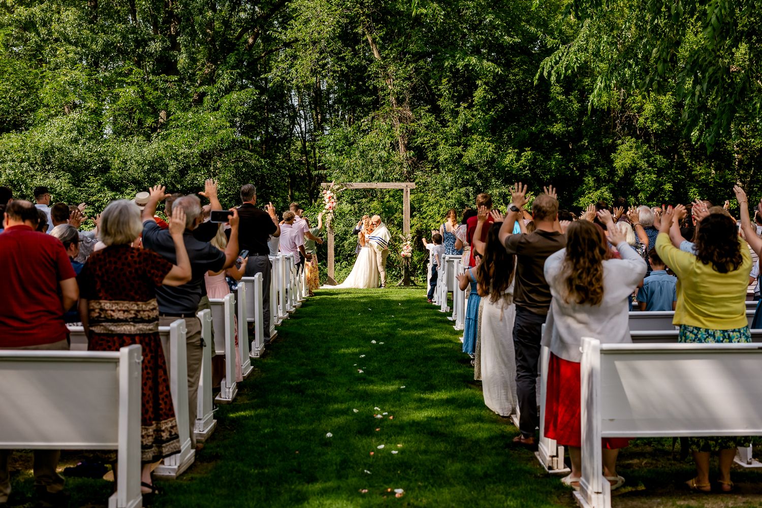 Outdoor wedding ceremony on green lawn aisle with guests cheering, framed by lush trees and a wooden arch.