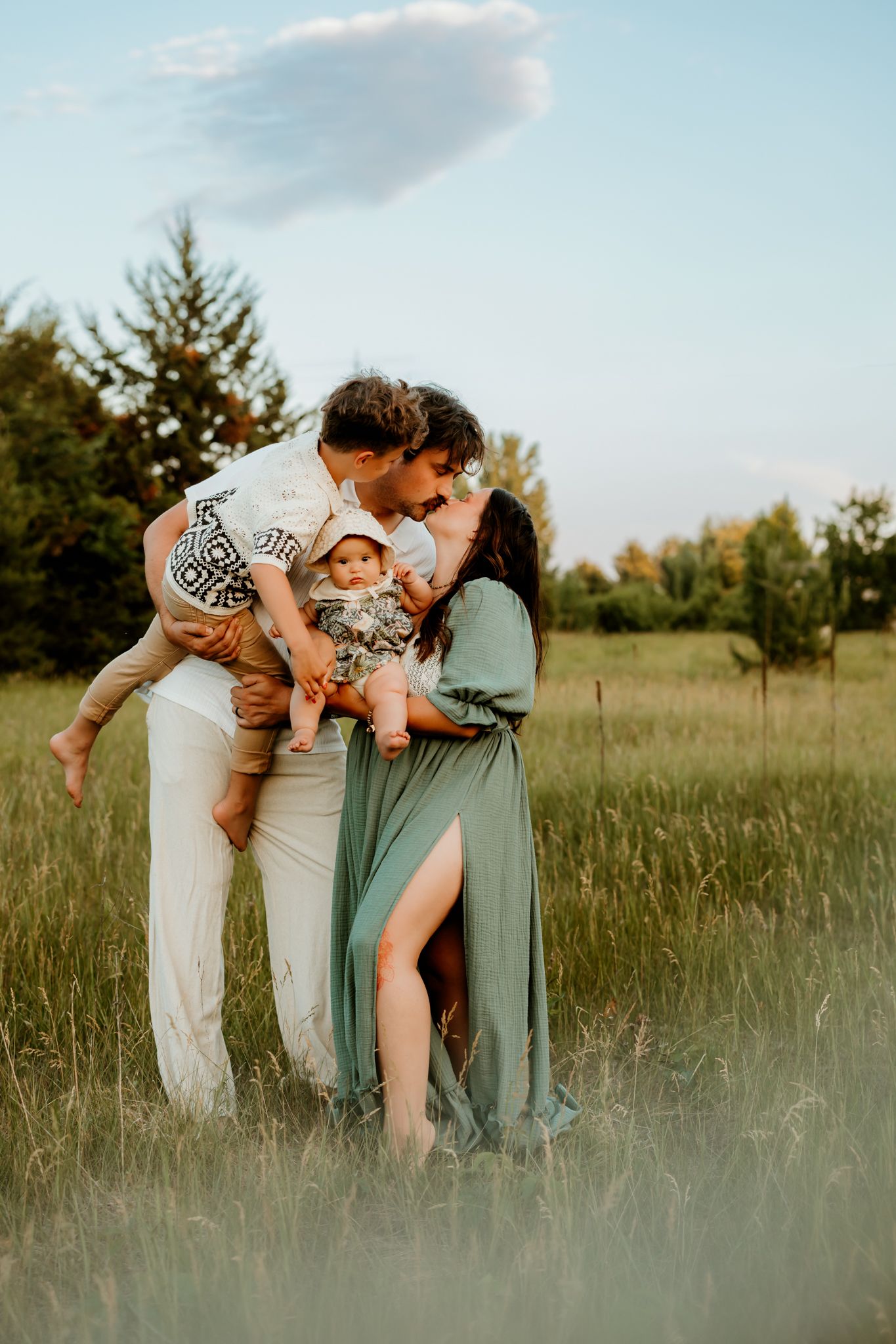 A couple kisses in a green field while holding their toddler and baby, wearing boho outfits on a sunny day.
