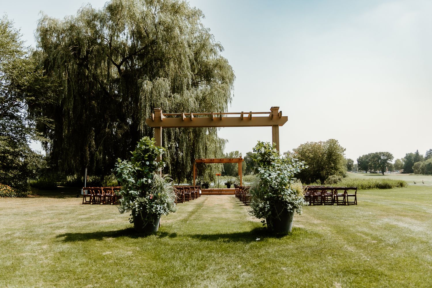 Outdoor wedding ceremony setup with wooden pergola, floral arrangements, and rustic benches on a lush green lawn.