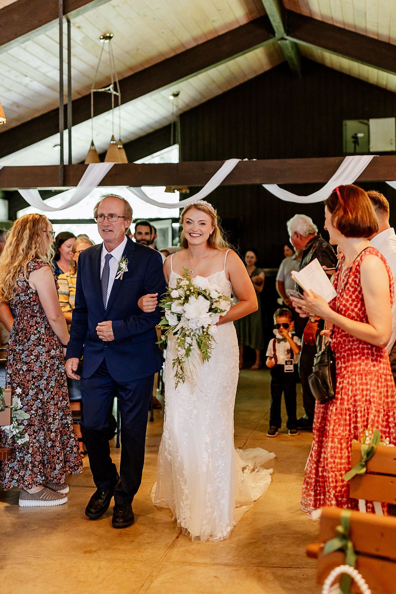 Bride in white lace gown walks down aisle with escort in navy suit, holding white floral bouquet in rustic barn venue.