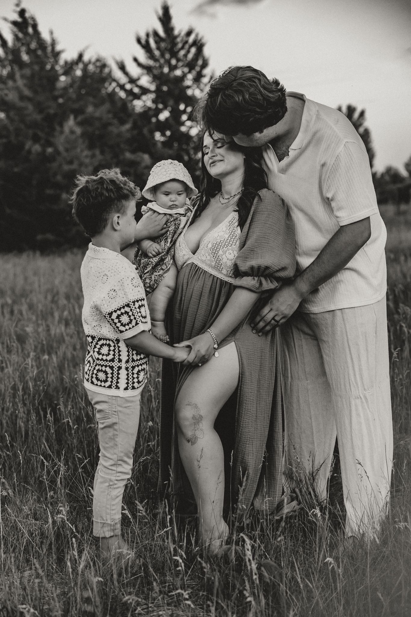 Black and white family maternity photo with pregnant mom, dad, and two children in a lush green field.