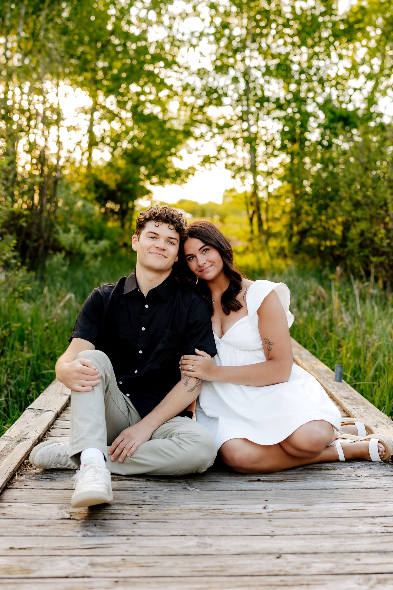 A happy couple sits together on a wooden boardwalk, smiling warmly, surrounded by green trees in golden sunlight.