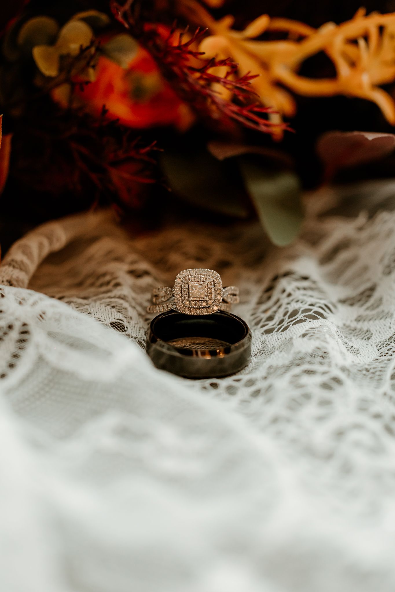 Diamond halo engagement ring resting on a black wedding band atop white lace, with autumn orange floral bouquet in the background.
