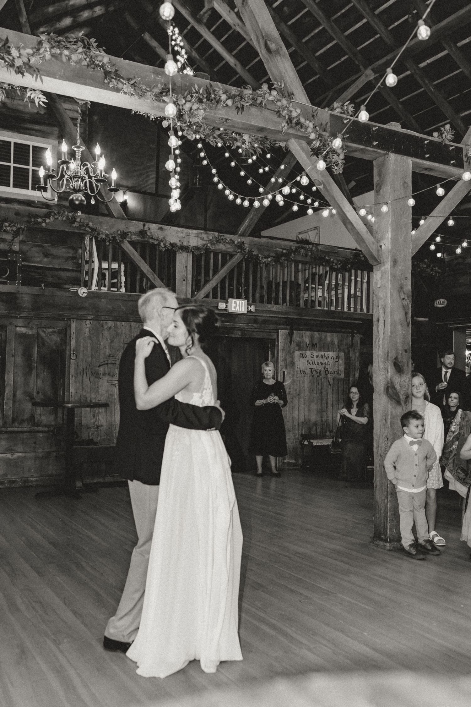 Bride in a flowing gown dances with an older man on the wooden floor of a rustic barn wedding venue strung with lights as guests watch.