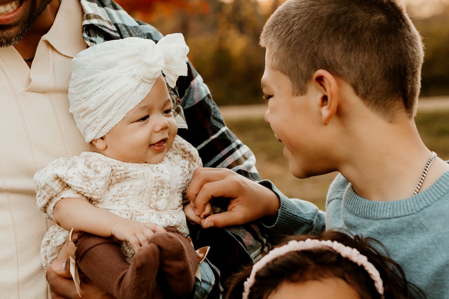 Smiling baby in a white knit turban and lace outfit being held by an adult while a boy in a blue sweater gently touches her hand outdoors.