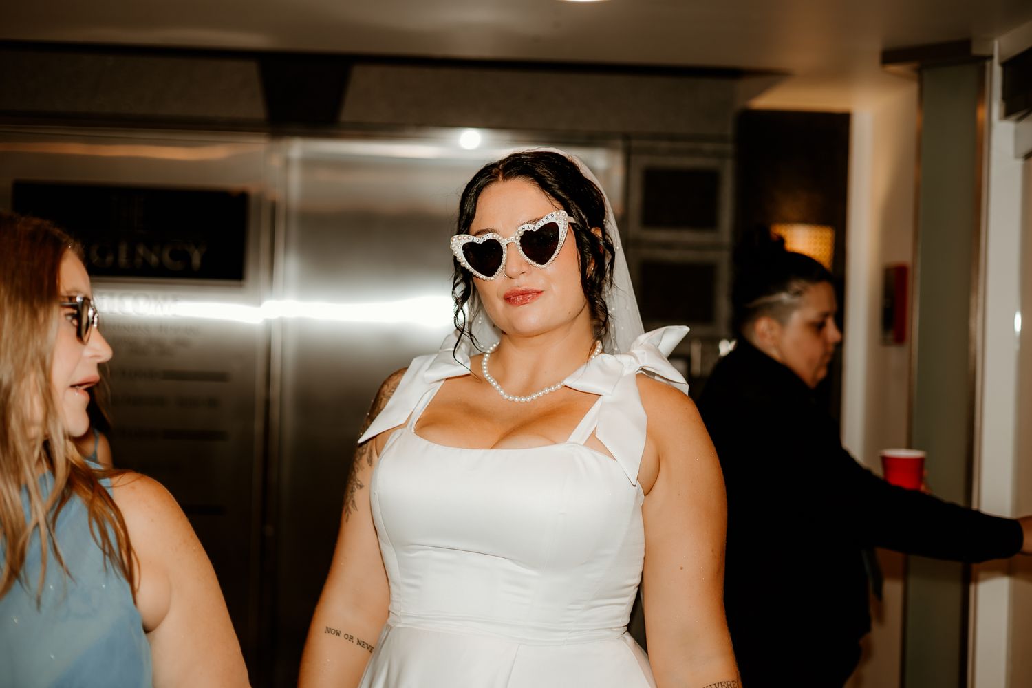 Bride in a white dress and heart-shaped white sunglasses stands in a hallway at a party, with a guest holding a red cup behind her.