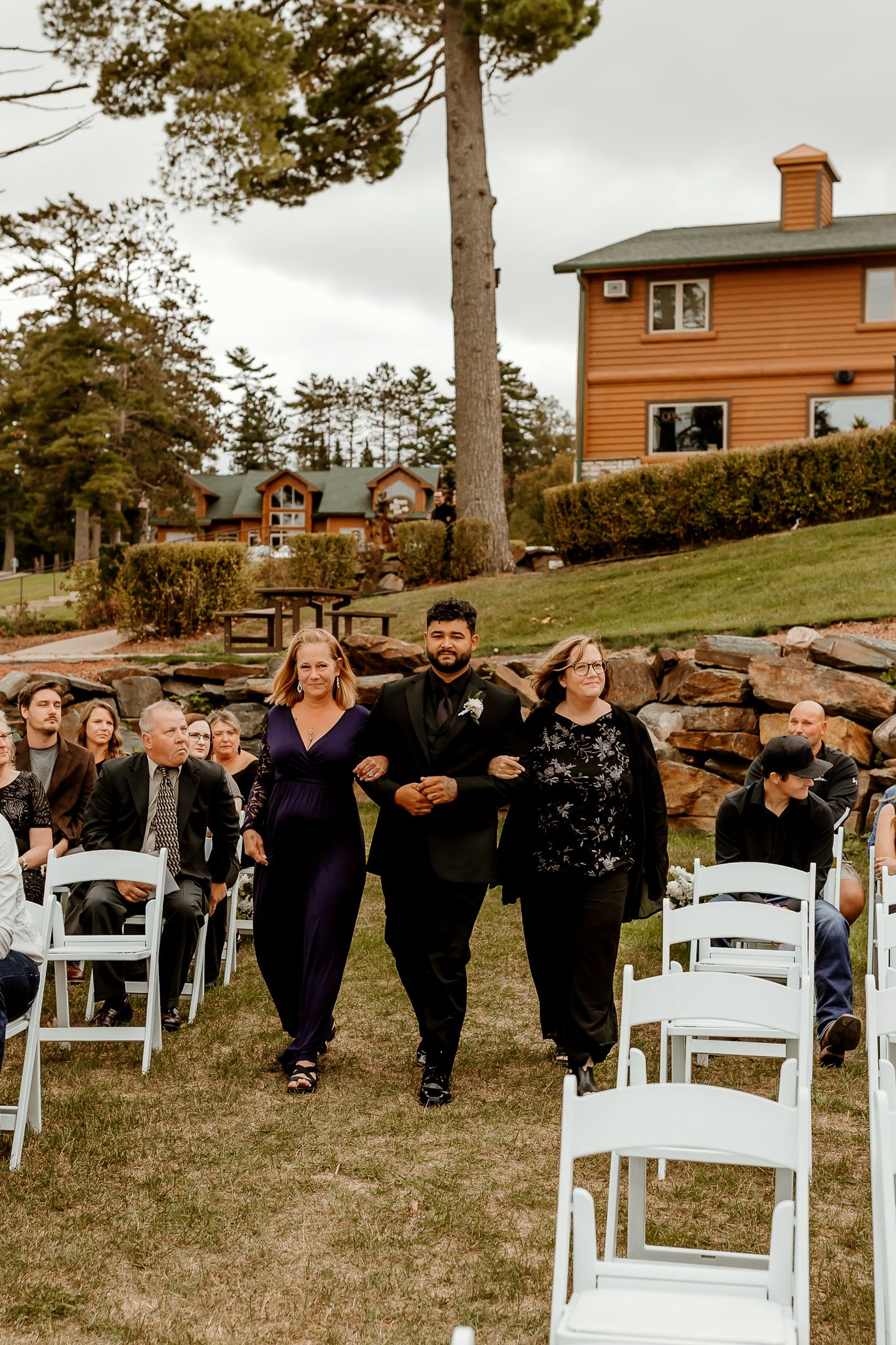 Three people walk arm in arm down the grass aisle past white chairs and seated guests at an outdoor lakeside wedding ceremony.