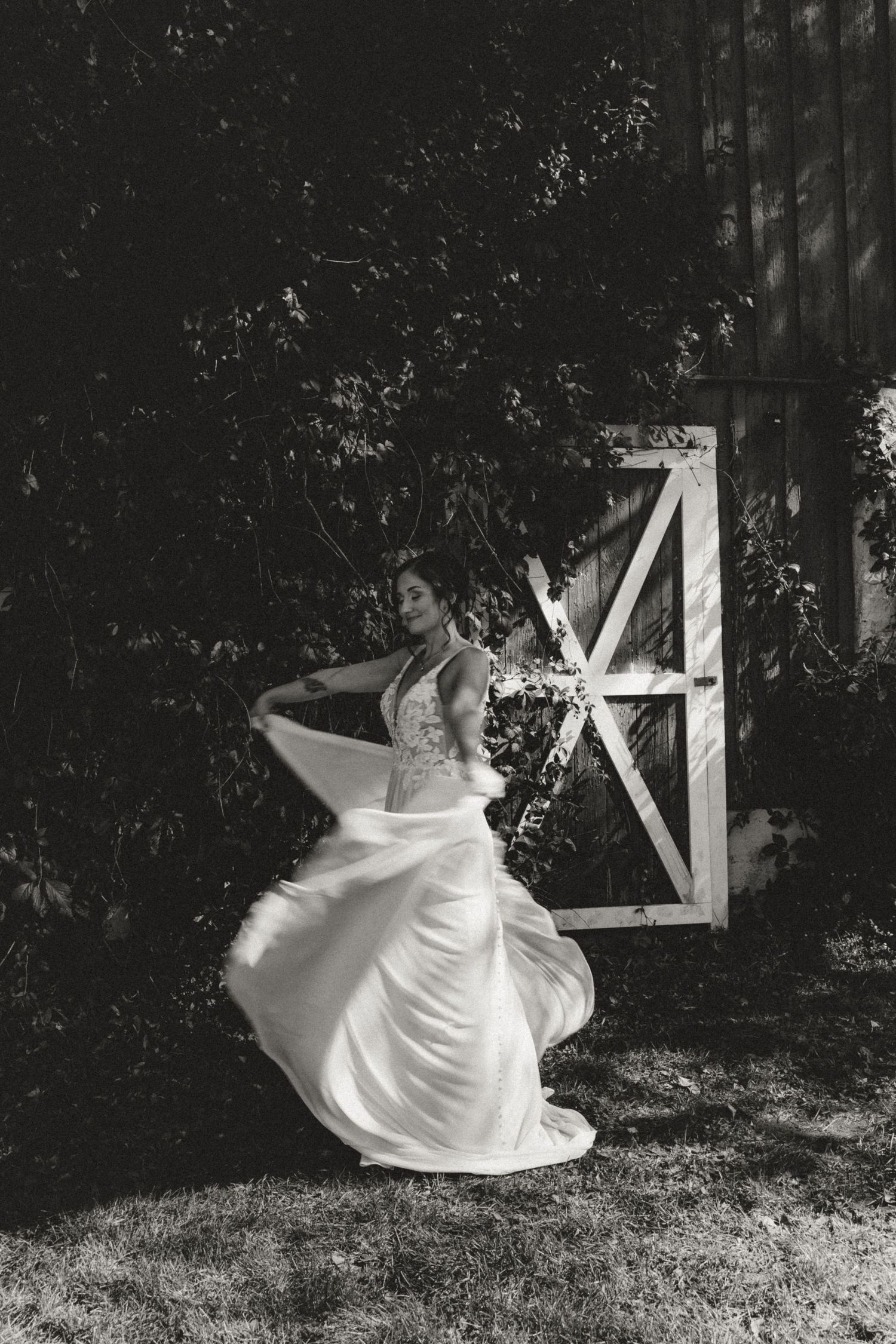 Black and white photo of a bride twirling in a flowing lace wedding dress beside a rustic barn door surrounded by lush greenery.