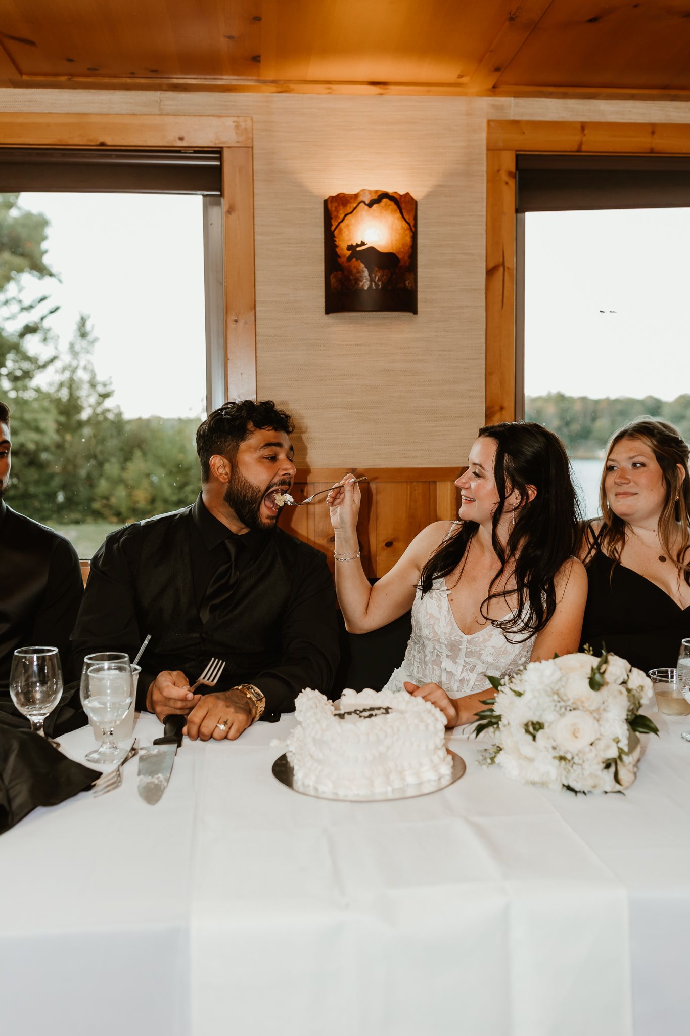 A bride feeds a bite of white wedding cake to a smiling groom at a reception table with a white floral bouquet beside a lakeside window.