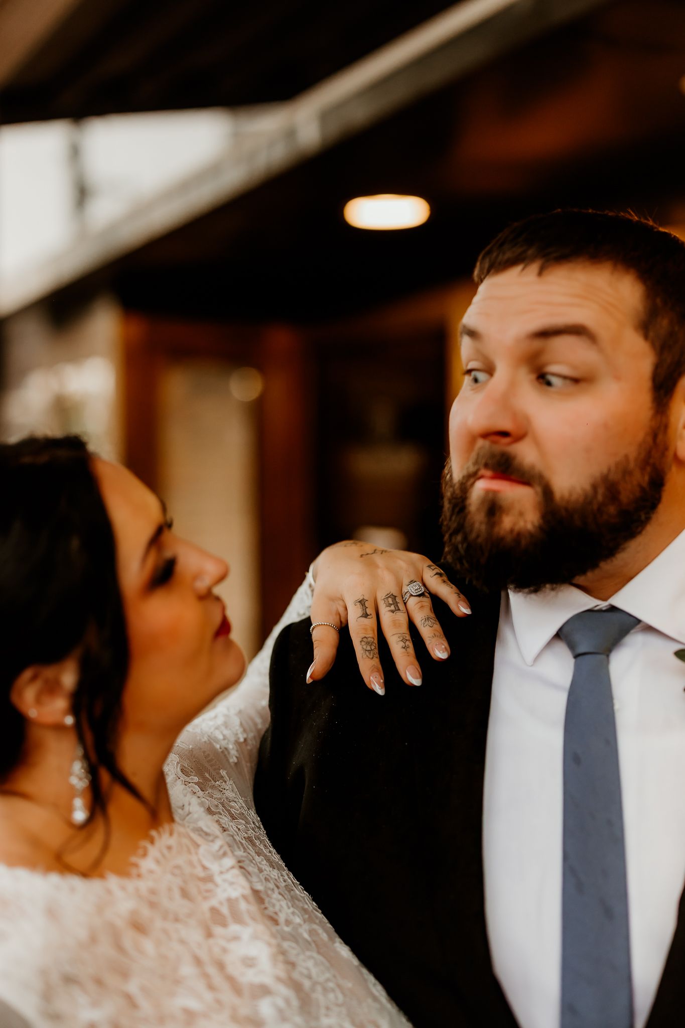 Bride in a white lace dress rests her tattooed hand on a bearded groom in a dark suit and gray tie during a wedding portrait.