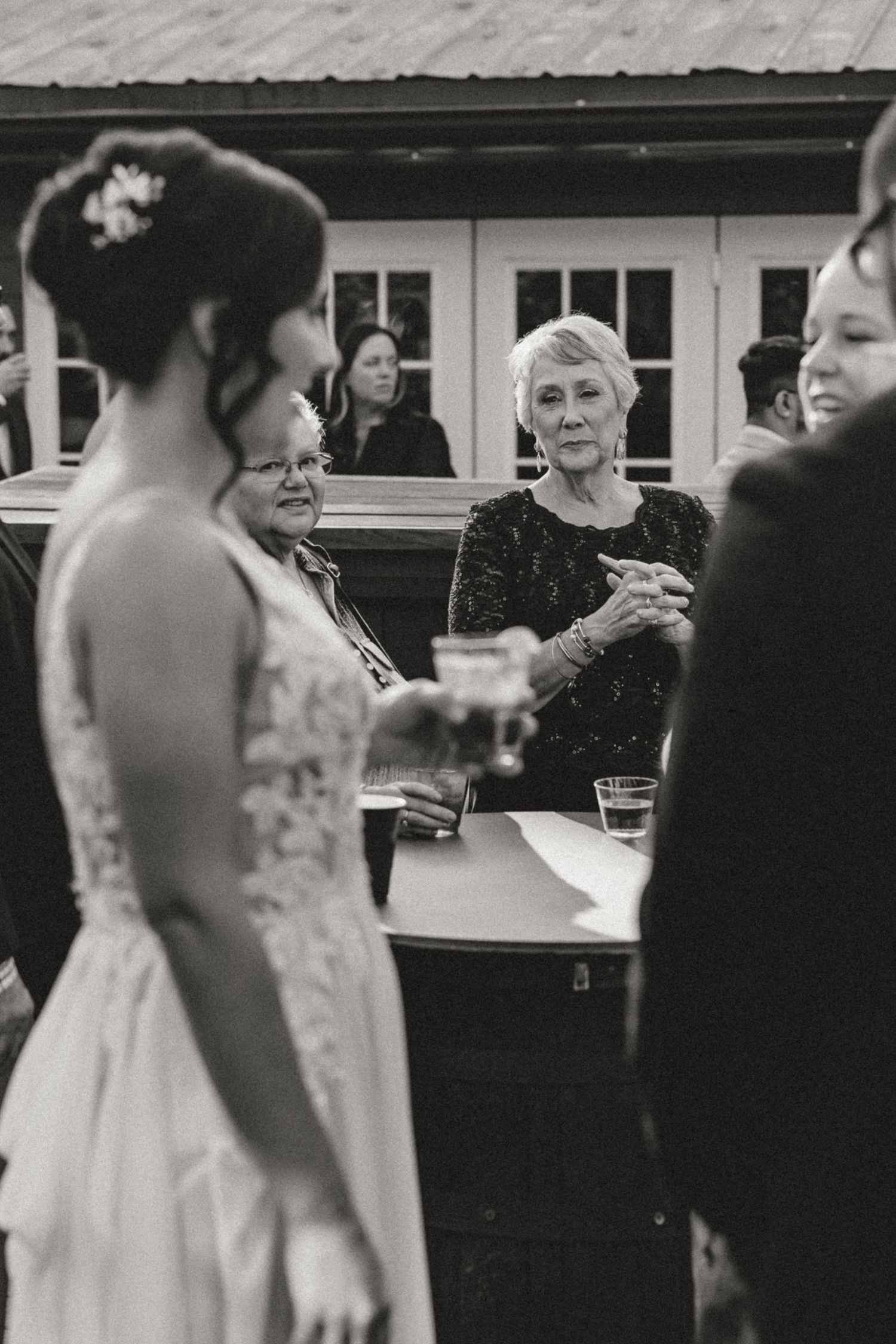 Black and white wedding photo of a bride in a lace gown holding a drink while an older woman watches from a table at an outdoor reception.