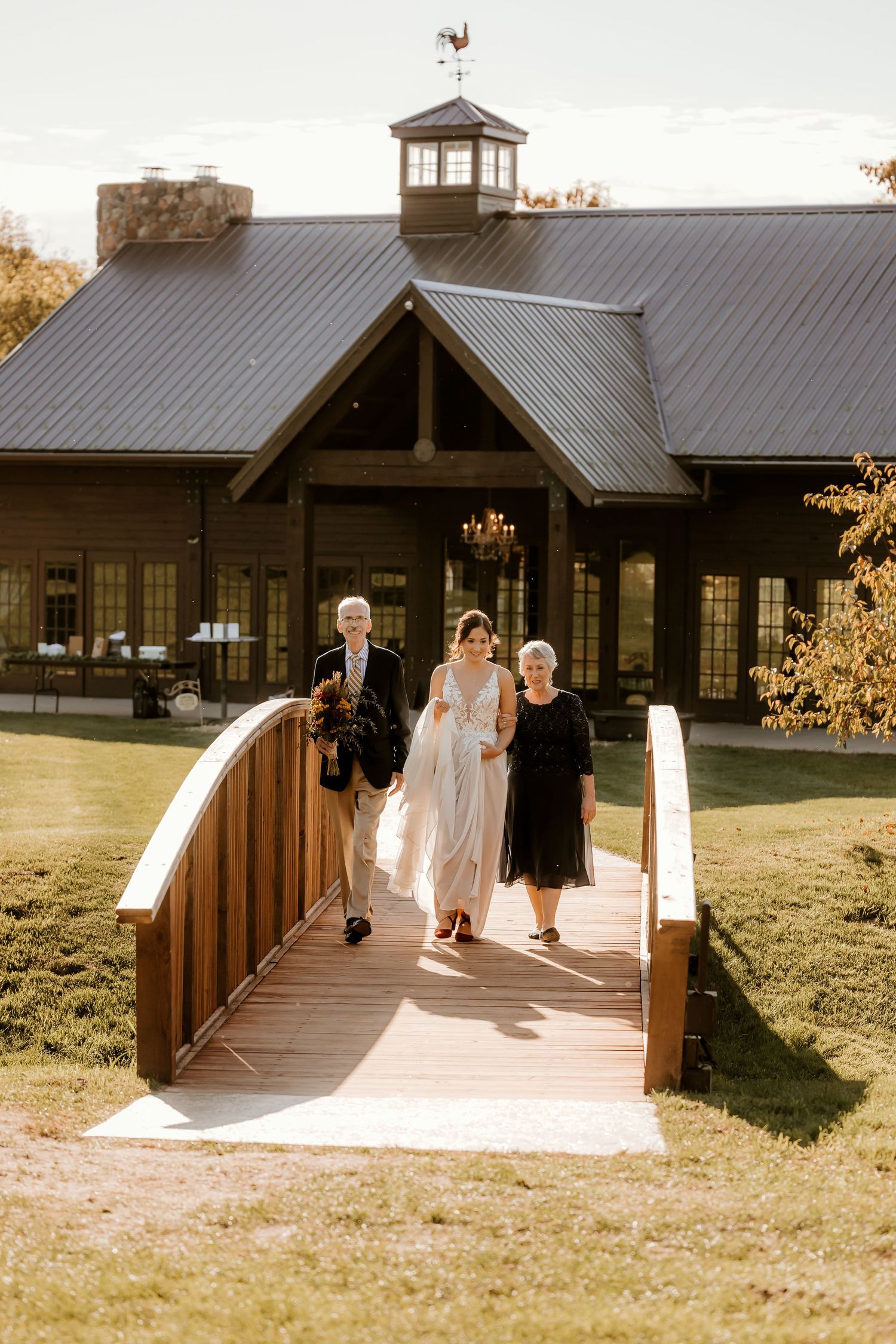 Bride in a flowing white gown walks across a wooden bridge with two companions toward a rustic barn-style wedding venue.