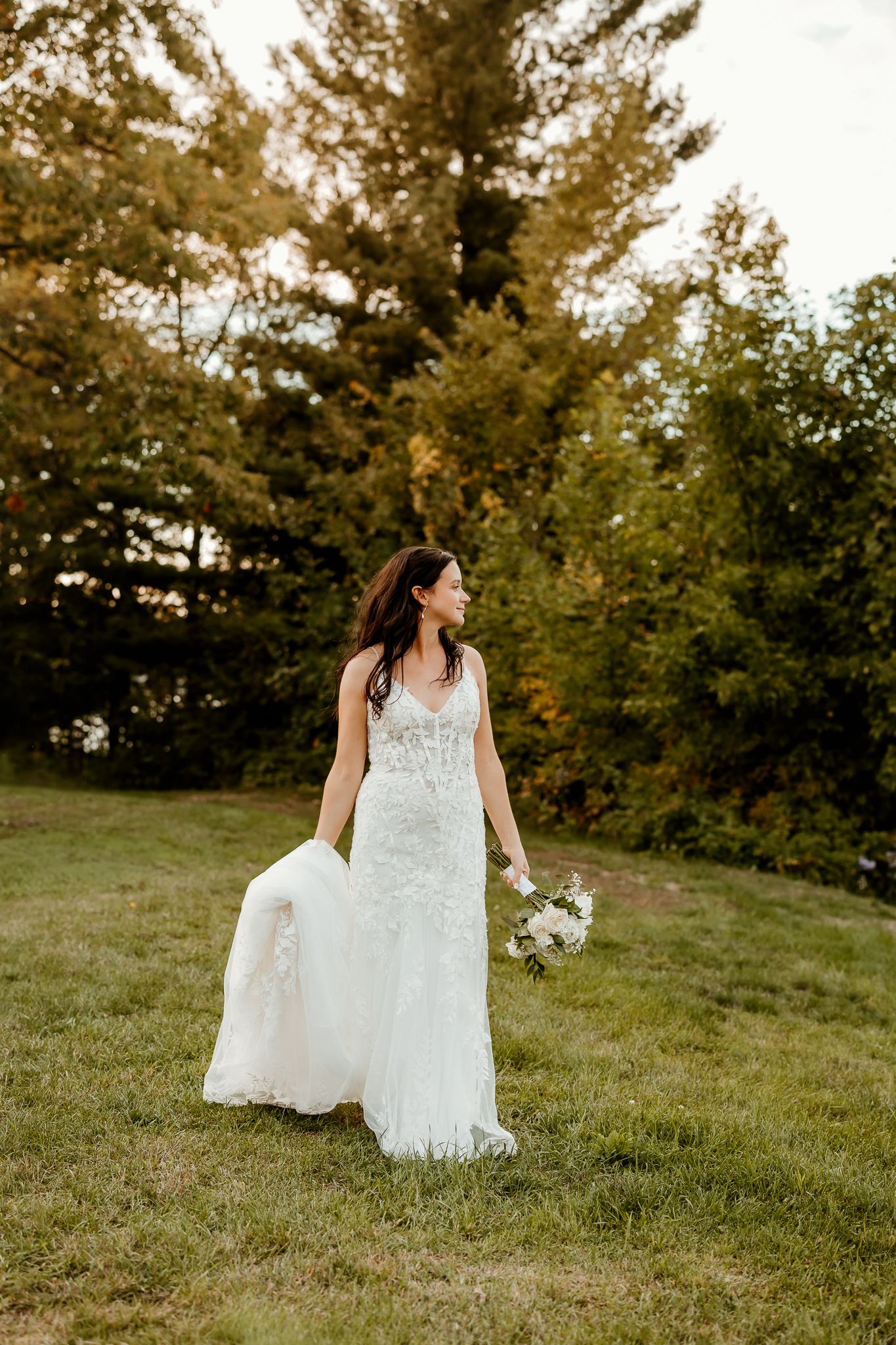 A bride in a lace wedding gown holds a white bouquet and looks over her shoulder while standing on a grassy hill with trees behind her.