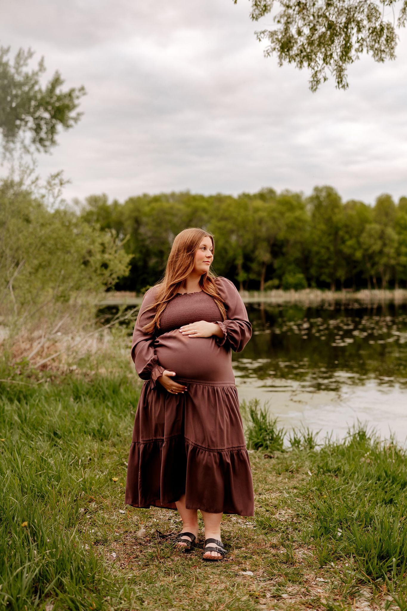 Pregnant woman in brown maxi dress cradles baby bump beside serene lake on cloudy day.