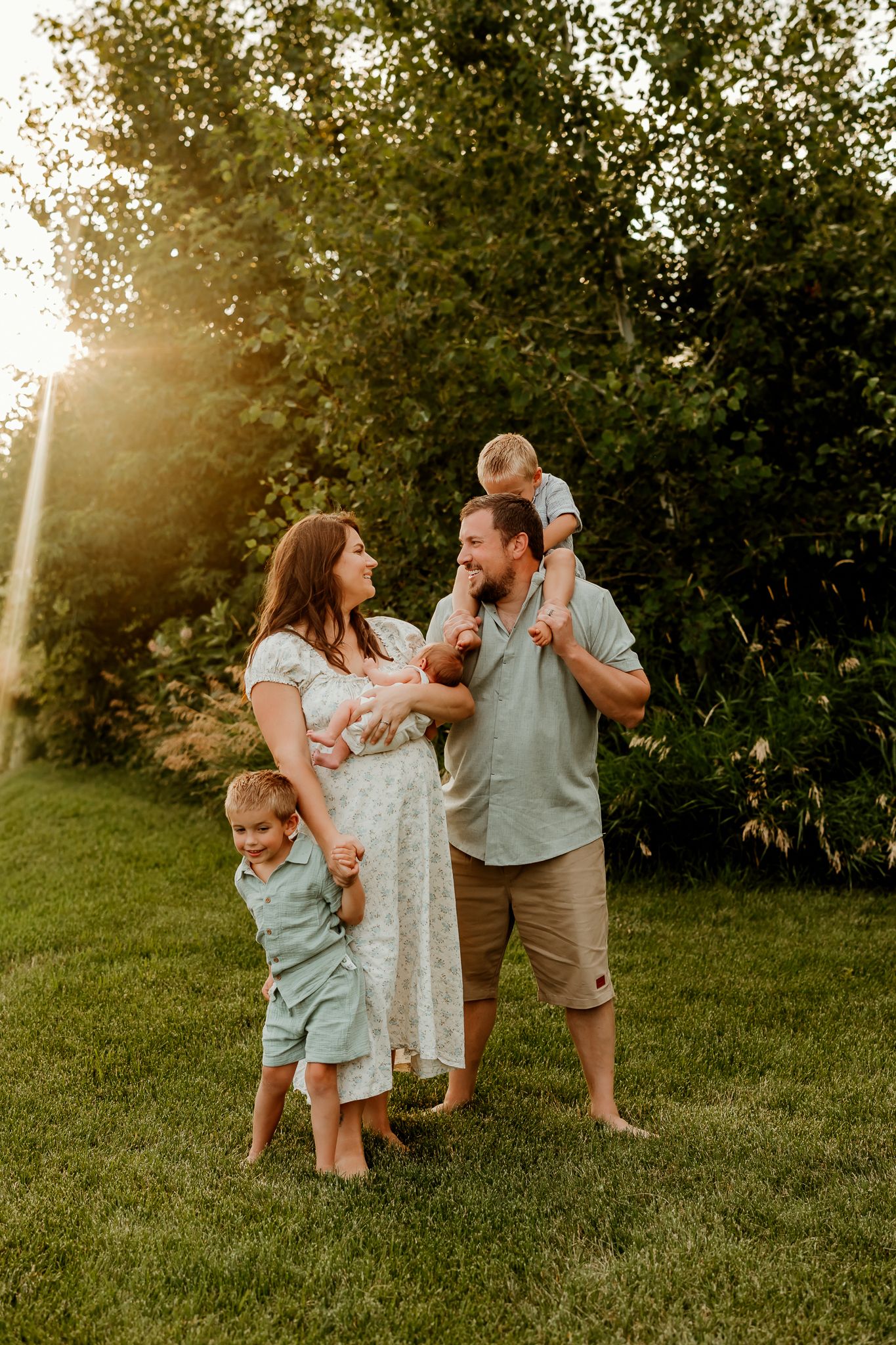 Happy family of five poses outdoors at golden hour, parents holding children on green grass with sunlit trees behind.