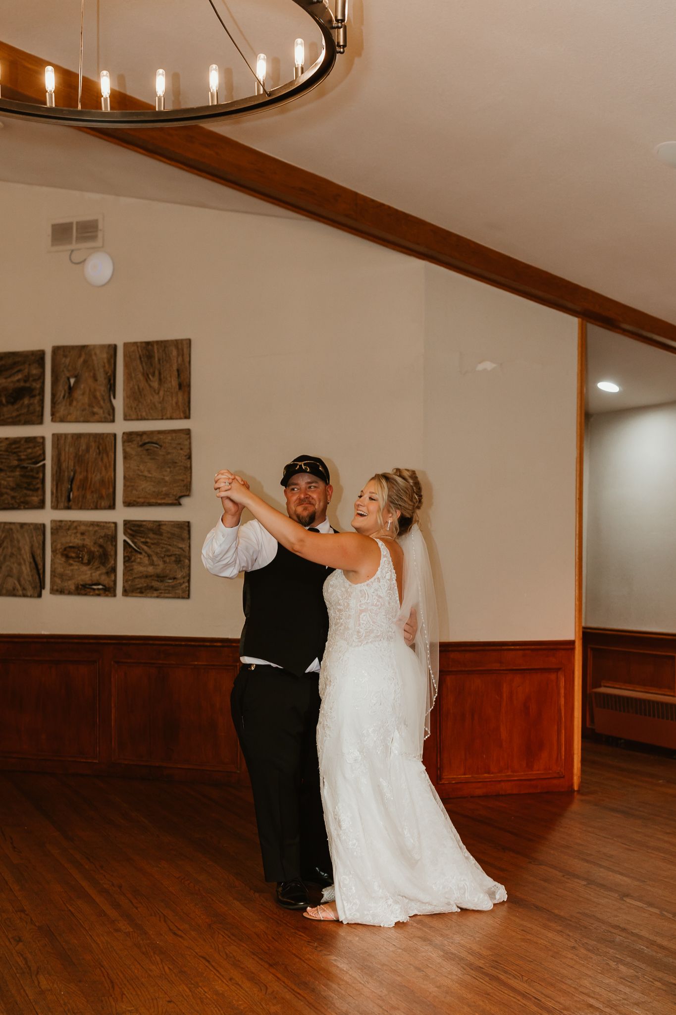 Bride and groom share their first dance in an elegant wood-paneled reception hall under a circular chandelier.