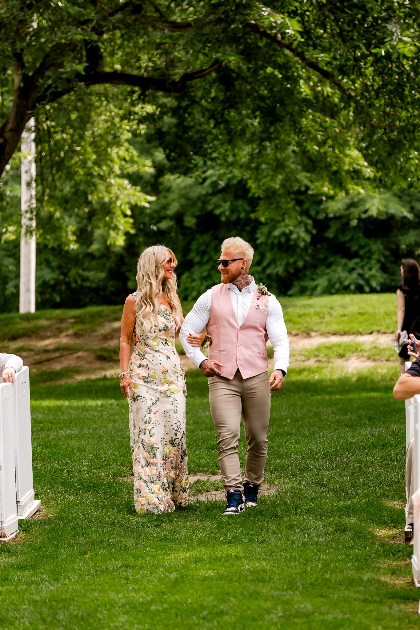Couple walks down outdoor wedding aisle, woman in floral dress, man in pink vest and khakis, lush green trees backdrop.
