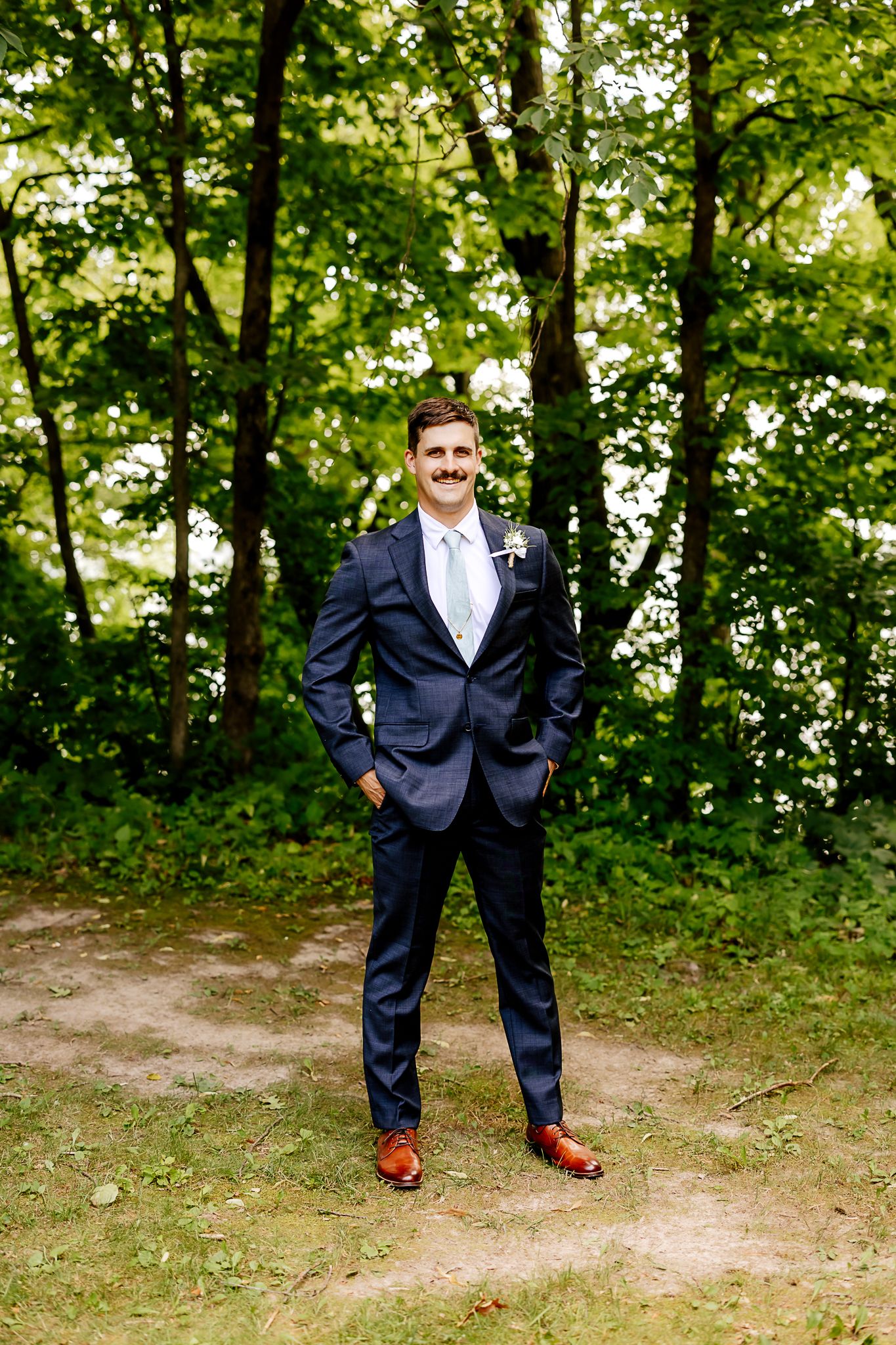 Groom in navy suit with boutonniere and brown shoes standing confidently in a lush green wooded outdoor setting.