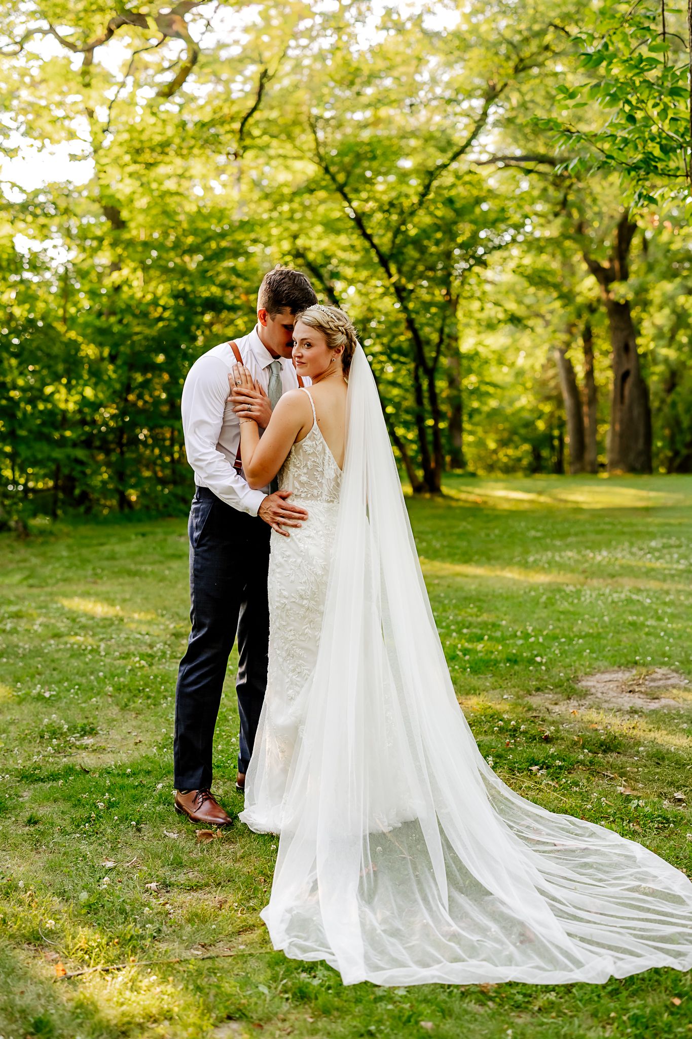 Bride in white gown and long veil embraced by groom in navy suit outdoors in lush green garden setting.