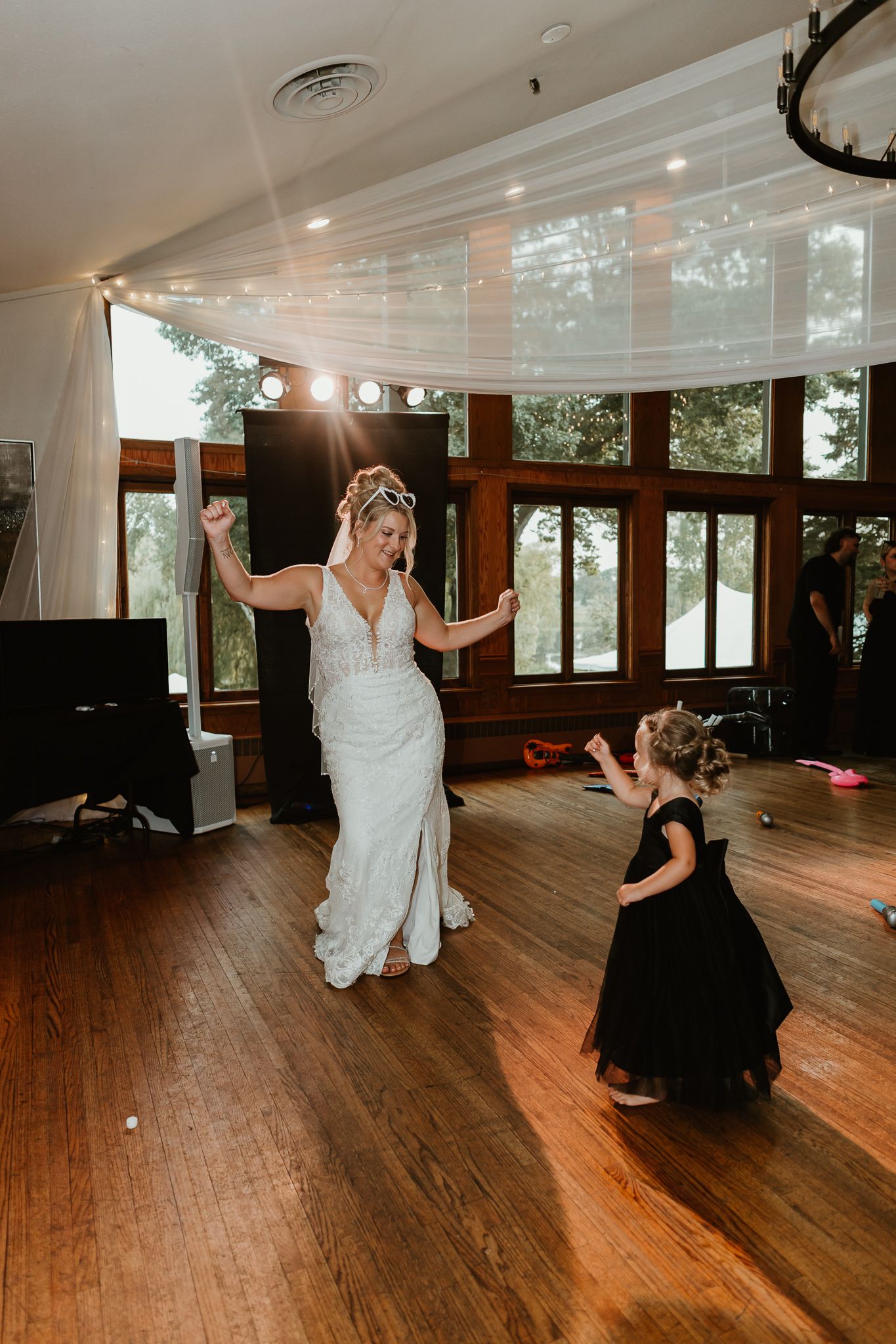 Bride in white gown dances joyfully with young flower girl in black dress on wooden dance floor at wedding reception.