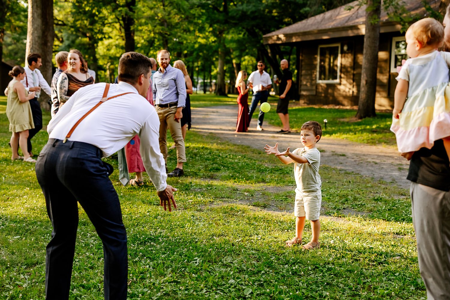 Man in suspenders plays with a toddler on a green lawn at an outdoor wedding reception.