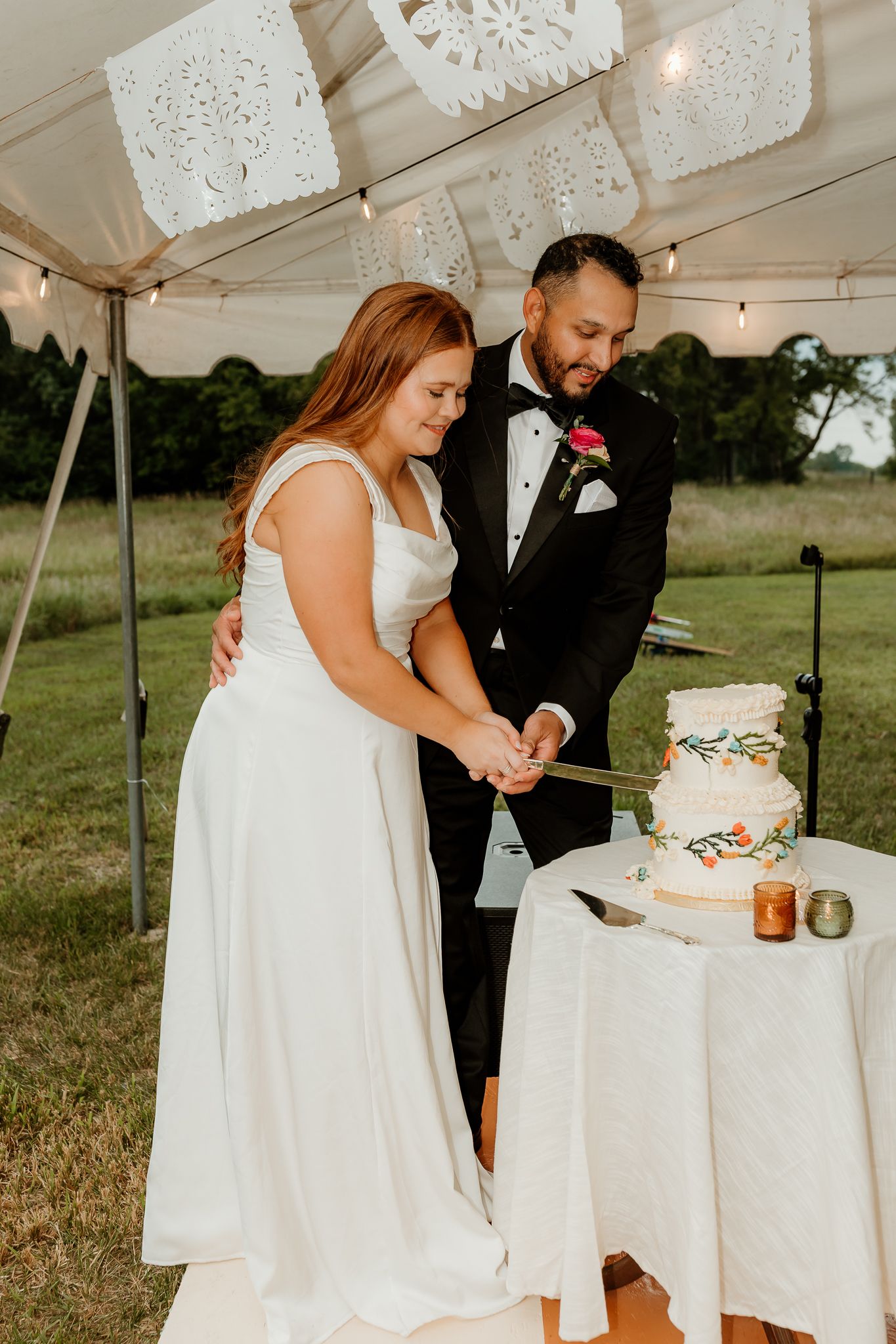 Bride and groom cutting their wedding cake under a tent decorated with paper banners at an outdoor reception.