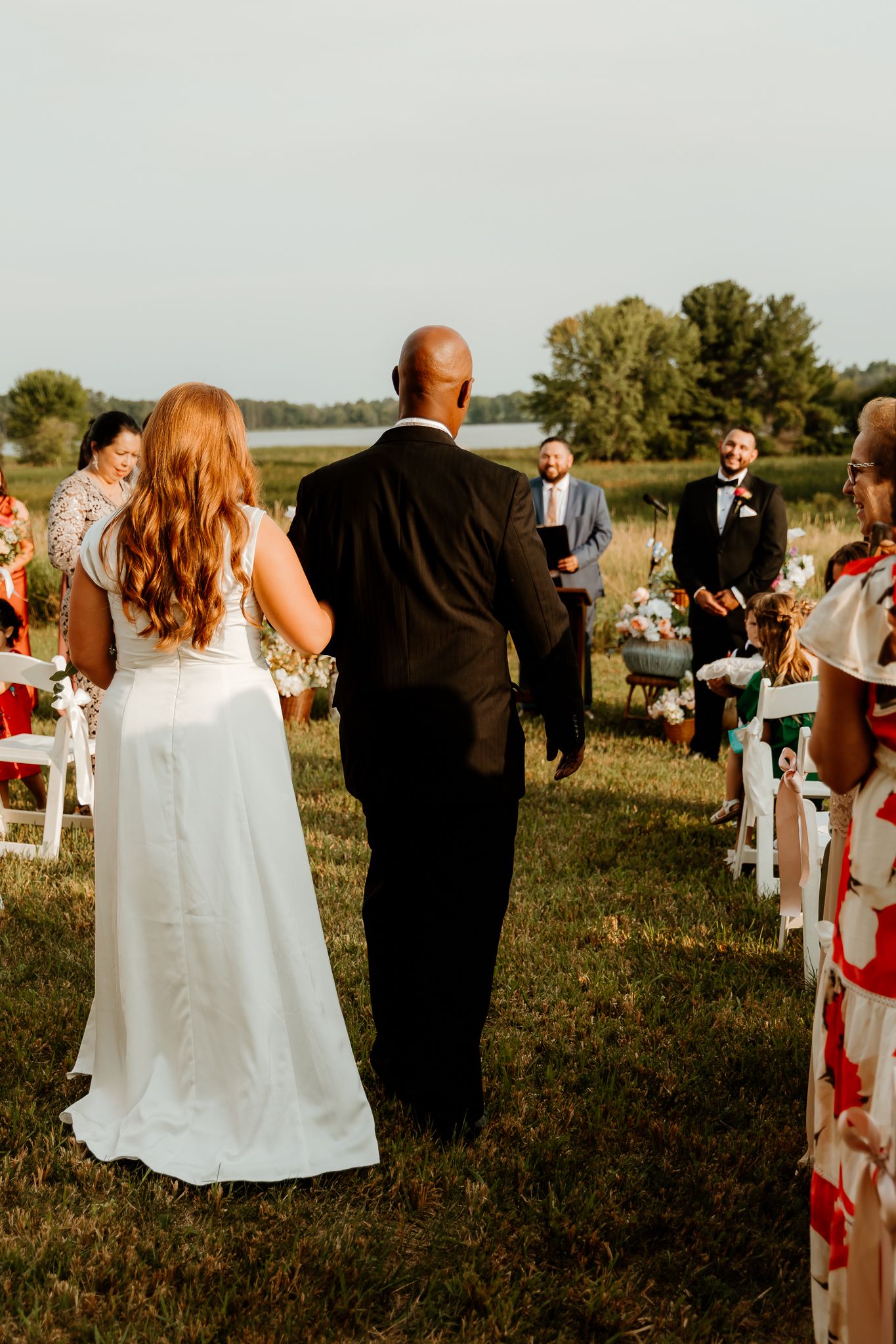 Bride in white gown walks down outdoor aisle with escort toward smiling groom at sunset wedding ceremony.