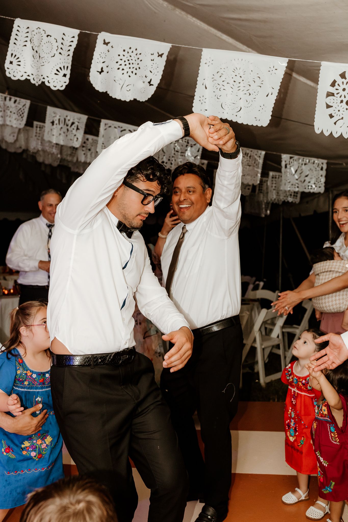 Two men dancing joyfully at a festive wedding reception decorated with papel picado banners.