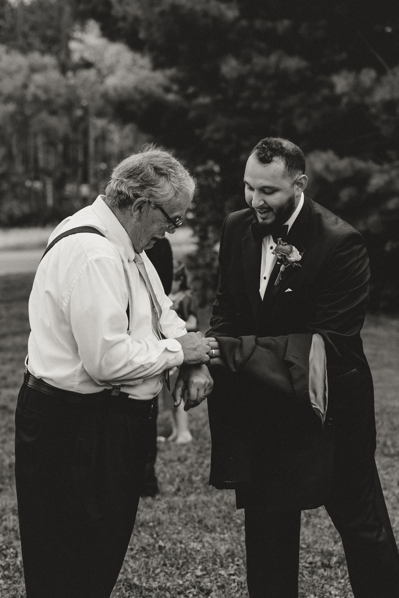 Groom in tuxedo helps older man with cufflinks outdoors at wedding in black and white photo.
