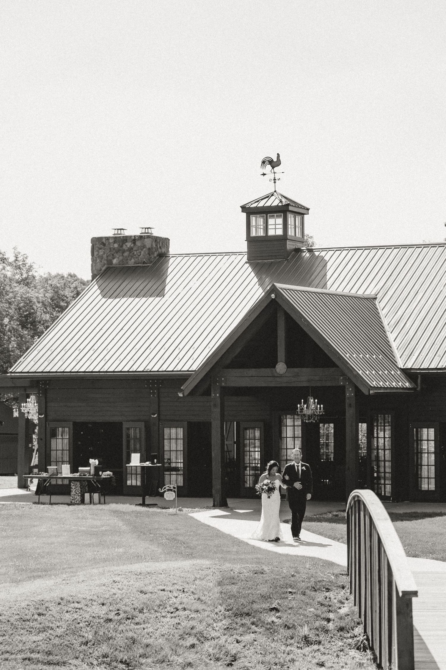 Black and white photo of a bride and groom walking outside a rustic barn wedding venue with a metal roof and weather vane.
