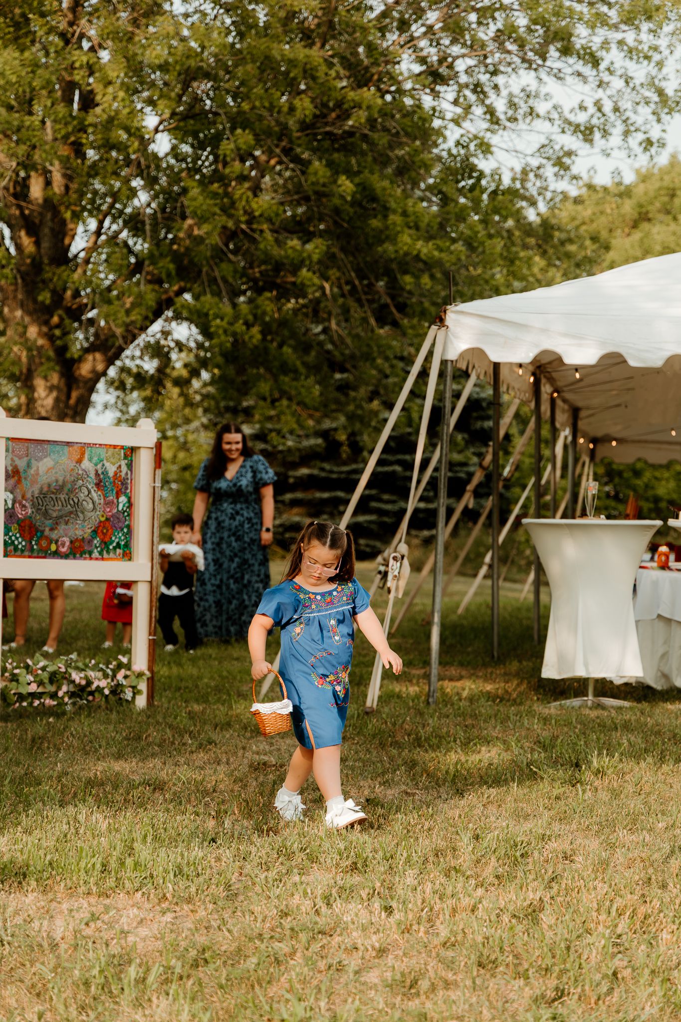 Young girl in denim overalls carrying a basket walks across grass at an outdoor event with a white tent.