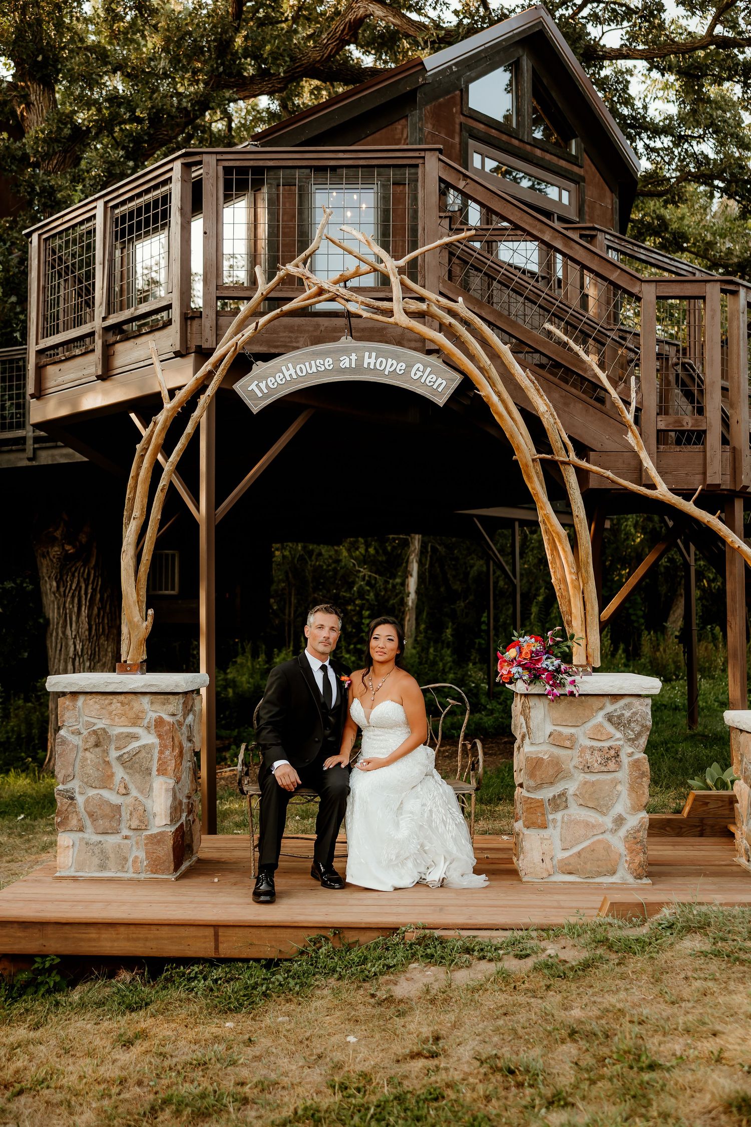 Bride and groom seated under rustic branch arch at TreeHouse at Hope Glen wedding venue with treehouse backdrop.