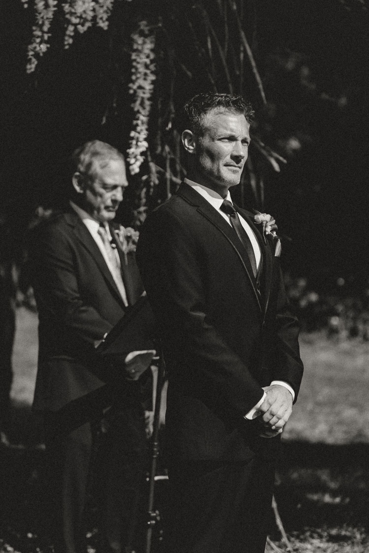 Groom in black suit smiling at altar during outdoor wedding ceremony, black and white photo.