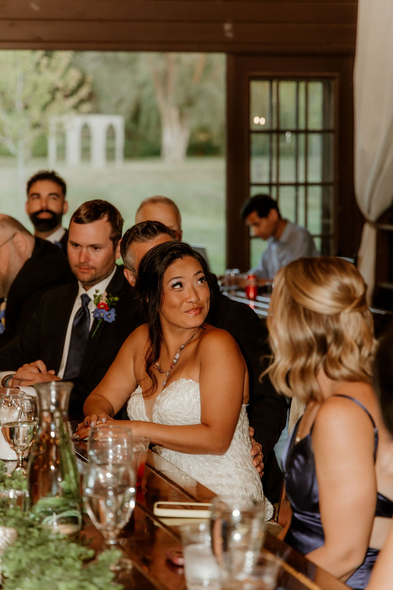 Bride in white lace gown sits at wedding reception table, surrounded by guests in formal attire, smiling warmly.
