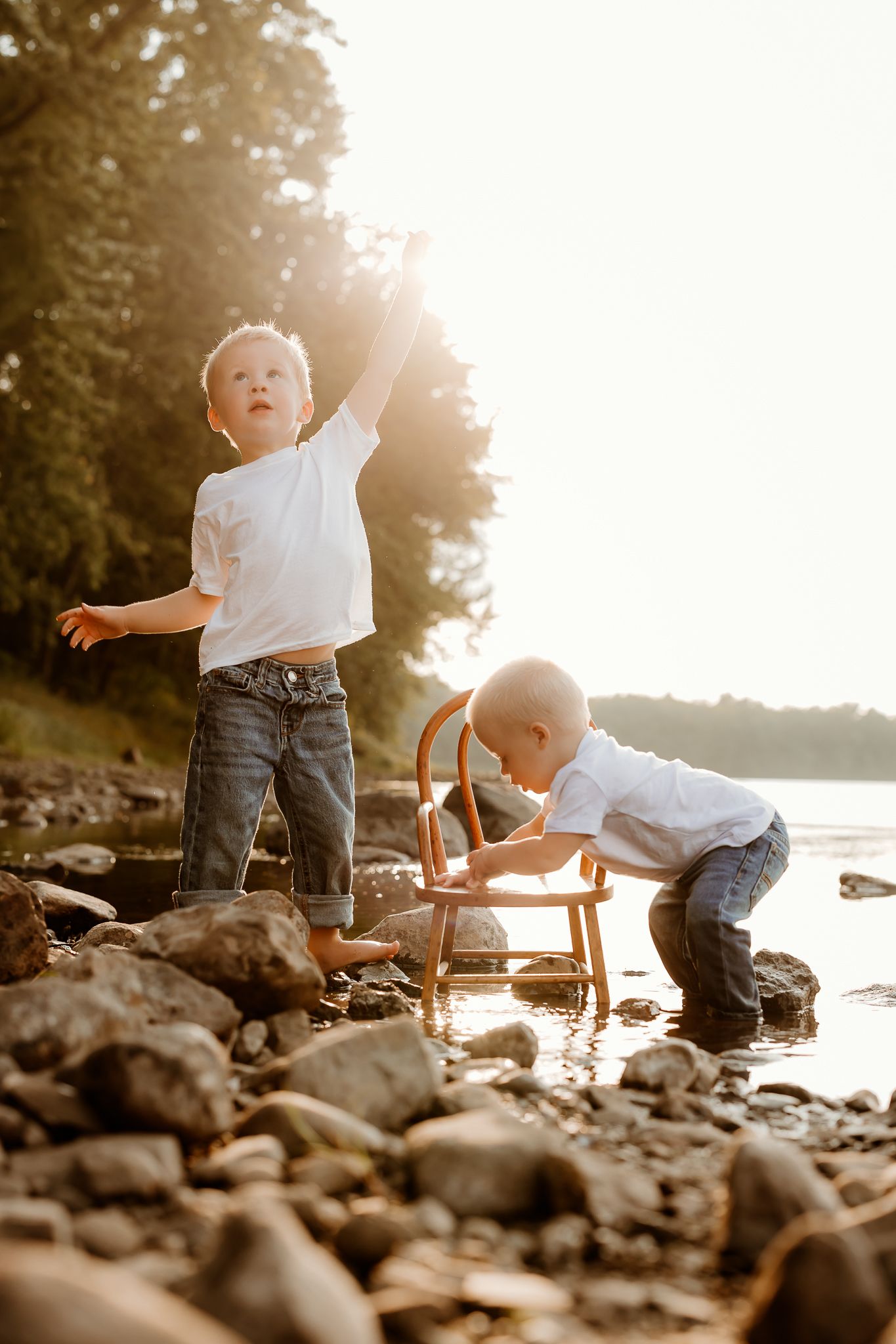 Two young boys playing on a rocky shoreline at golden hour, one reaching up while the other examines a small wooden chair.