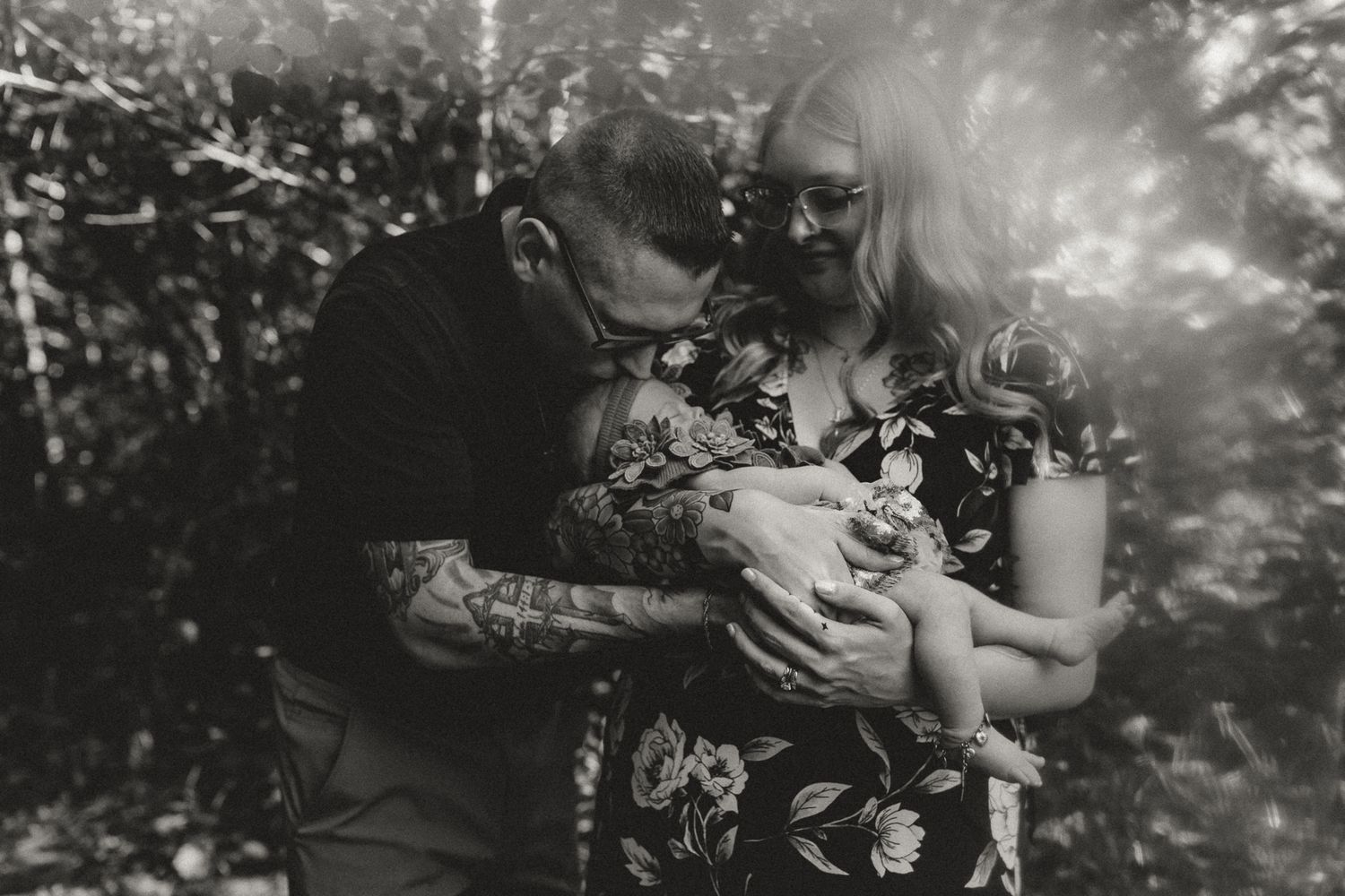 Black and white photo of tattooed couple kissing newborn baby outdoors among lush green trees.