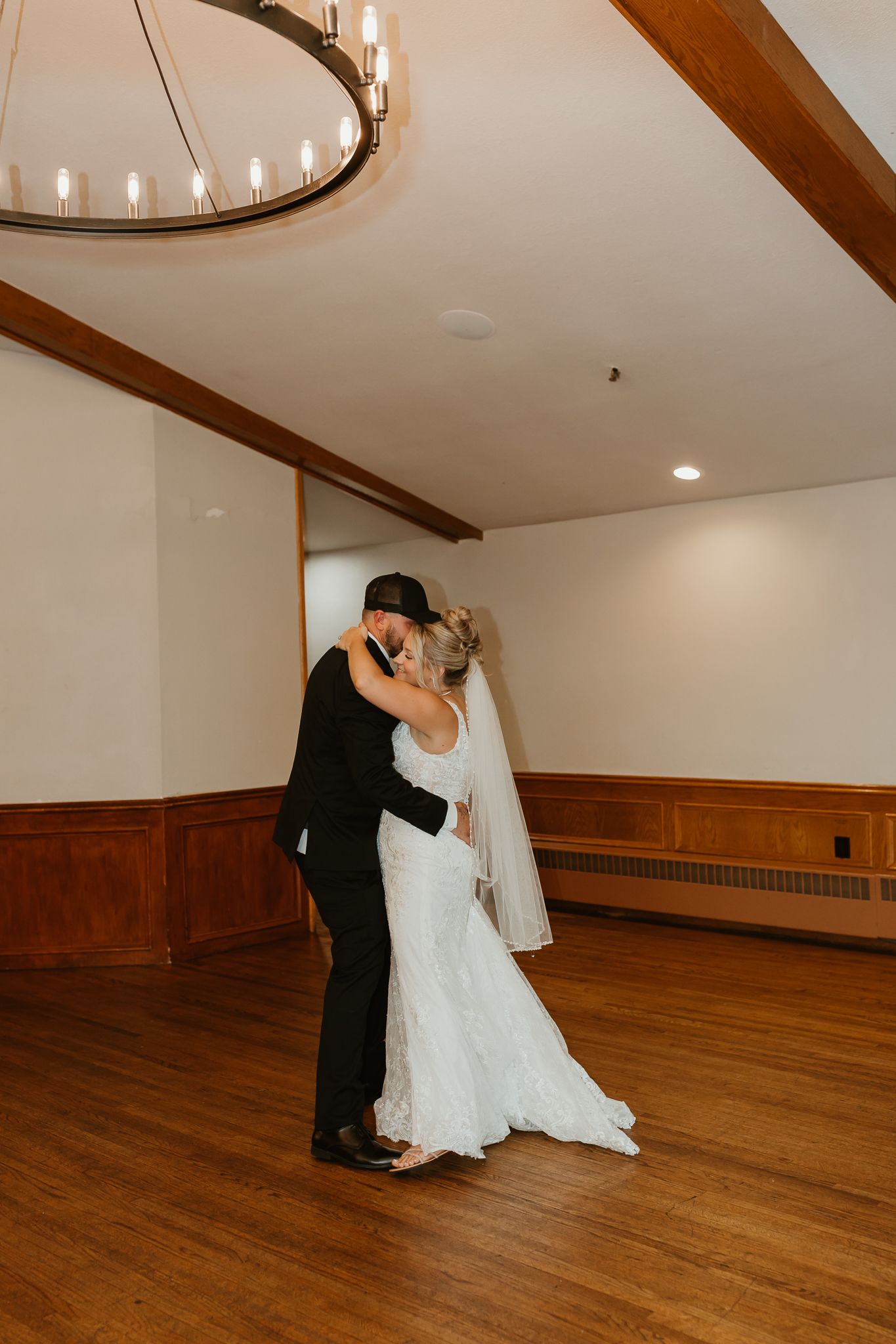 Bride and groom share first dance kiss in elegant wood-paneled ballroom with chandelier overhead.