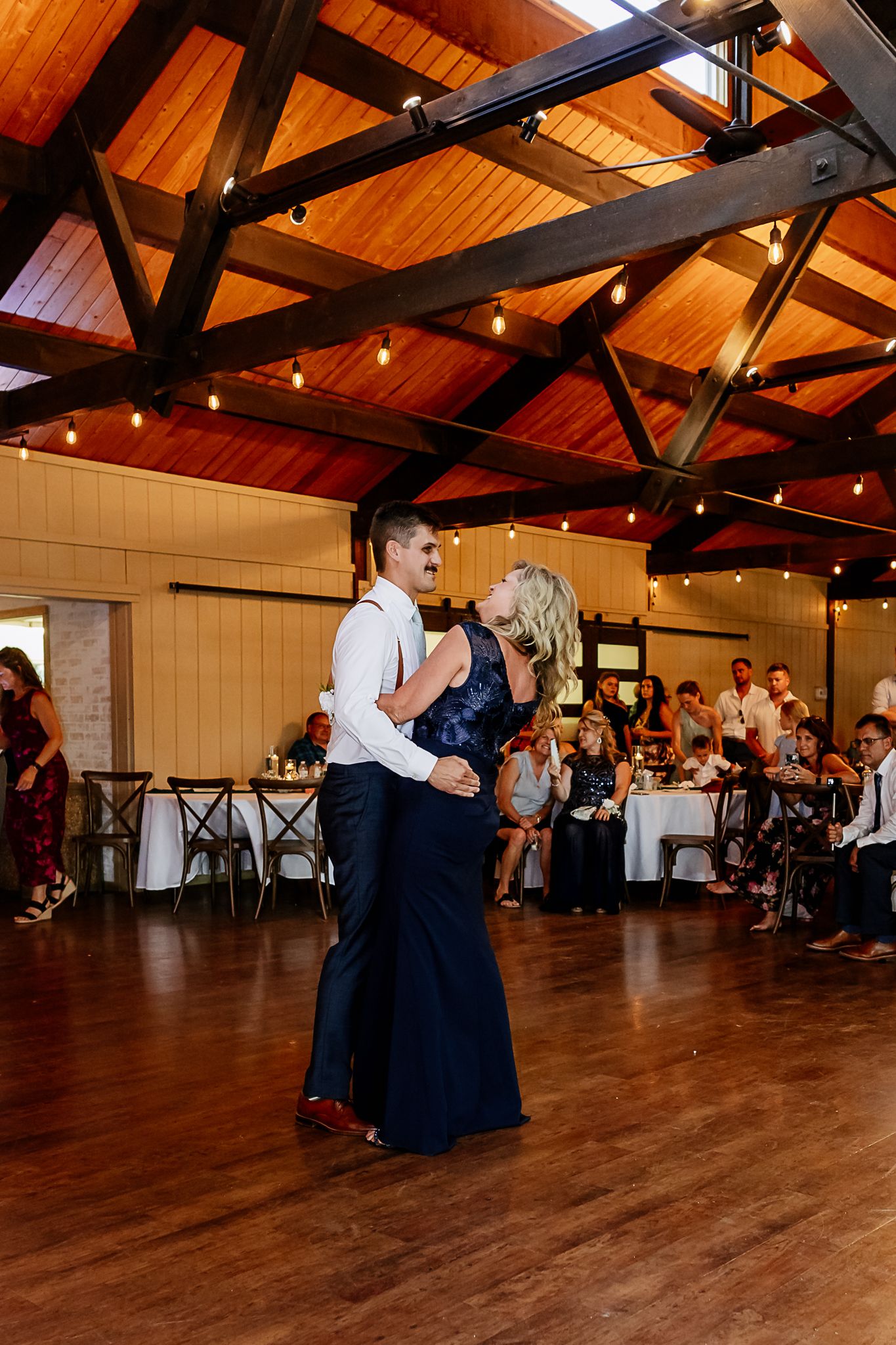 Couple sharing a romantic mother-son dance at a rustic wedding reception with warm string lights and wood beam ceiling.