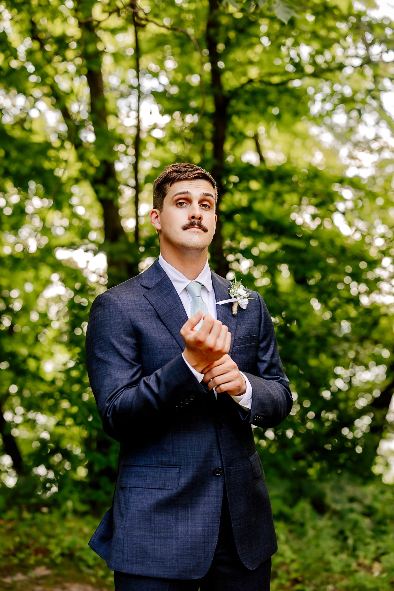 Groom in navy suit with mustache adjusting cufflinks outdoors, wearing boutonniere and light blue tie.