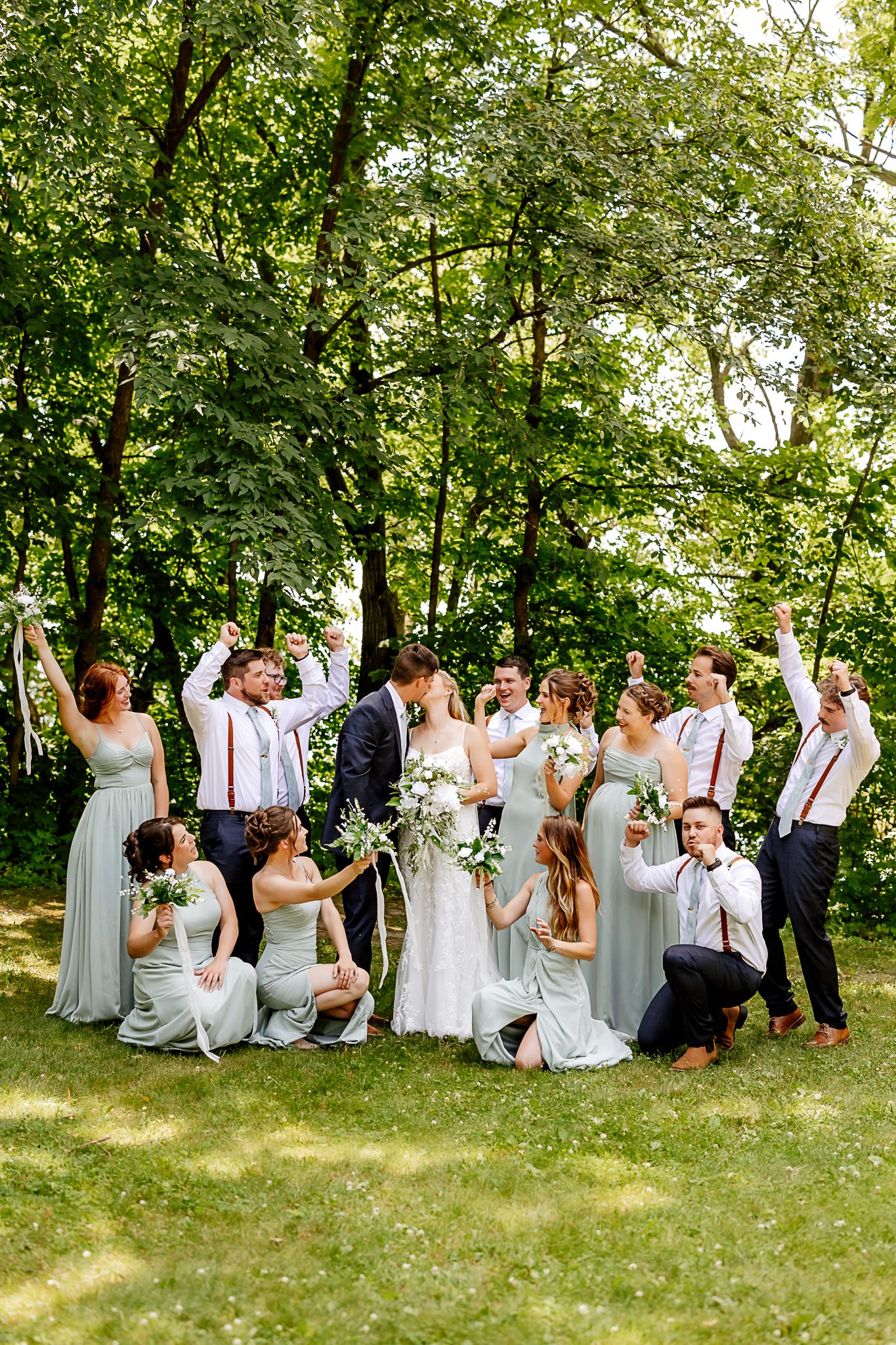 Wedding party celebrating outdoors in lush green park, bridesmaids in sage green dresses with groomsmen in suspenders.