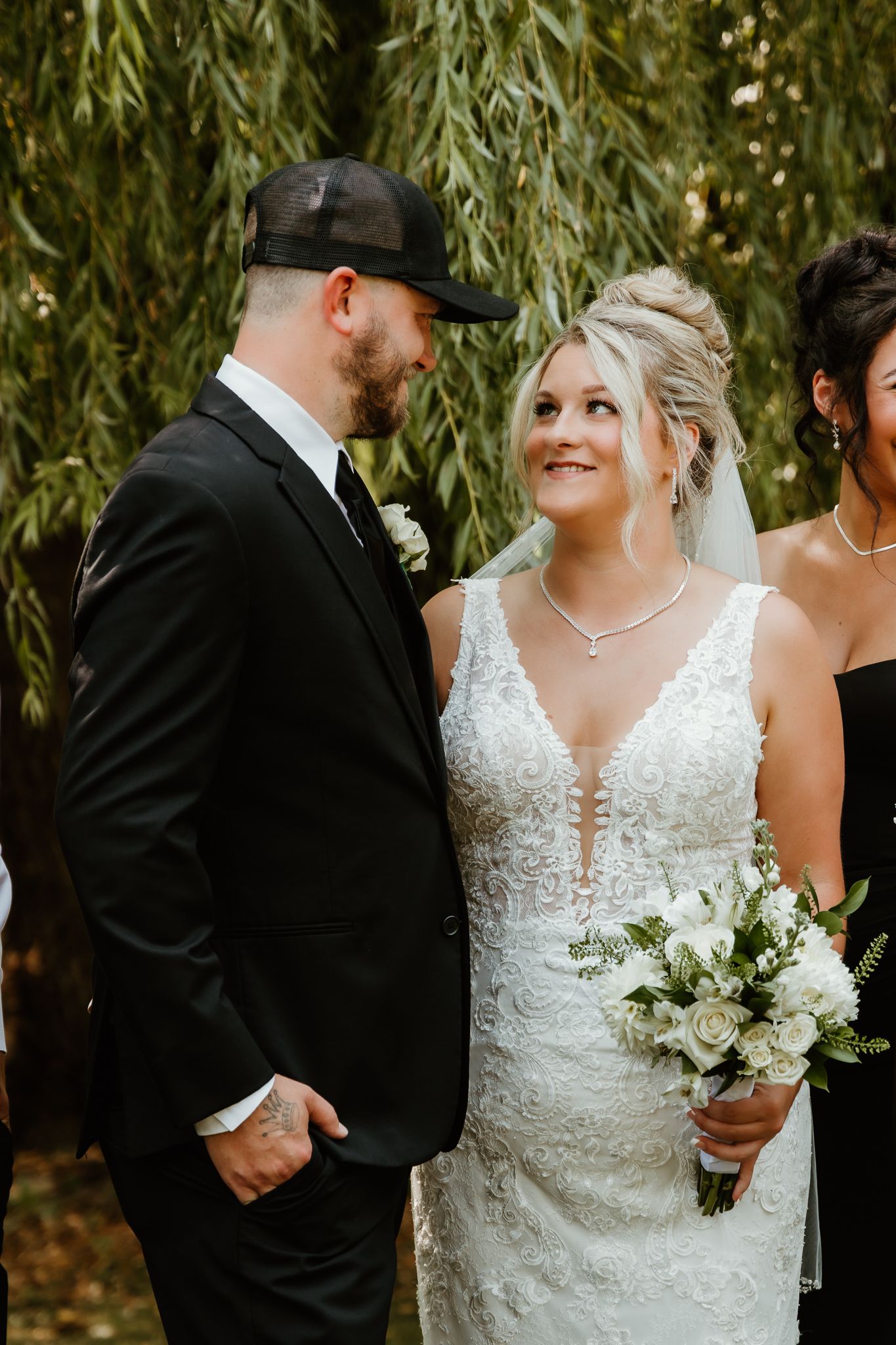 Bride in lace gown holding white bouquet smiles at groom in black suit and cap under willow tree.