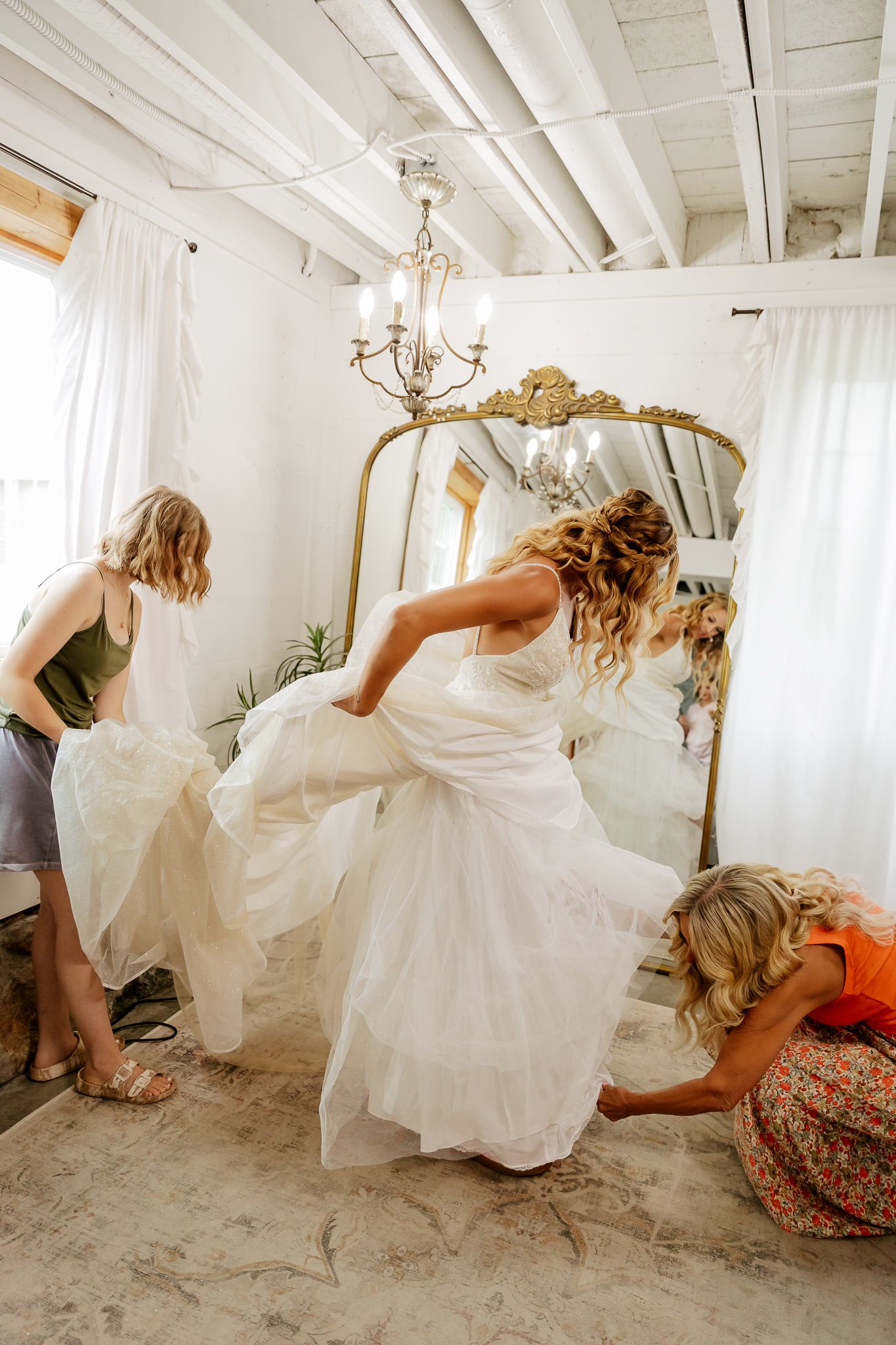 Bride in white gown gets help from bridesmaids in bright room with ornate gold mirror.