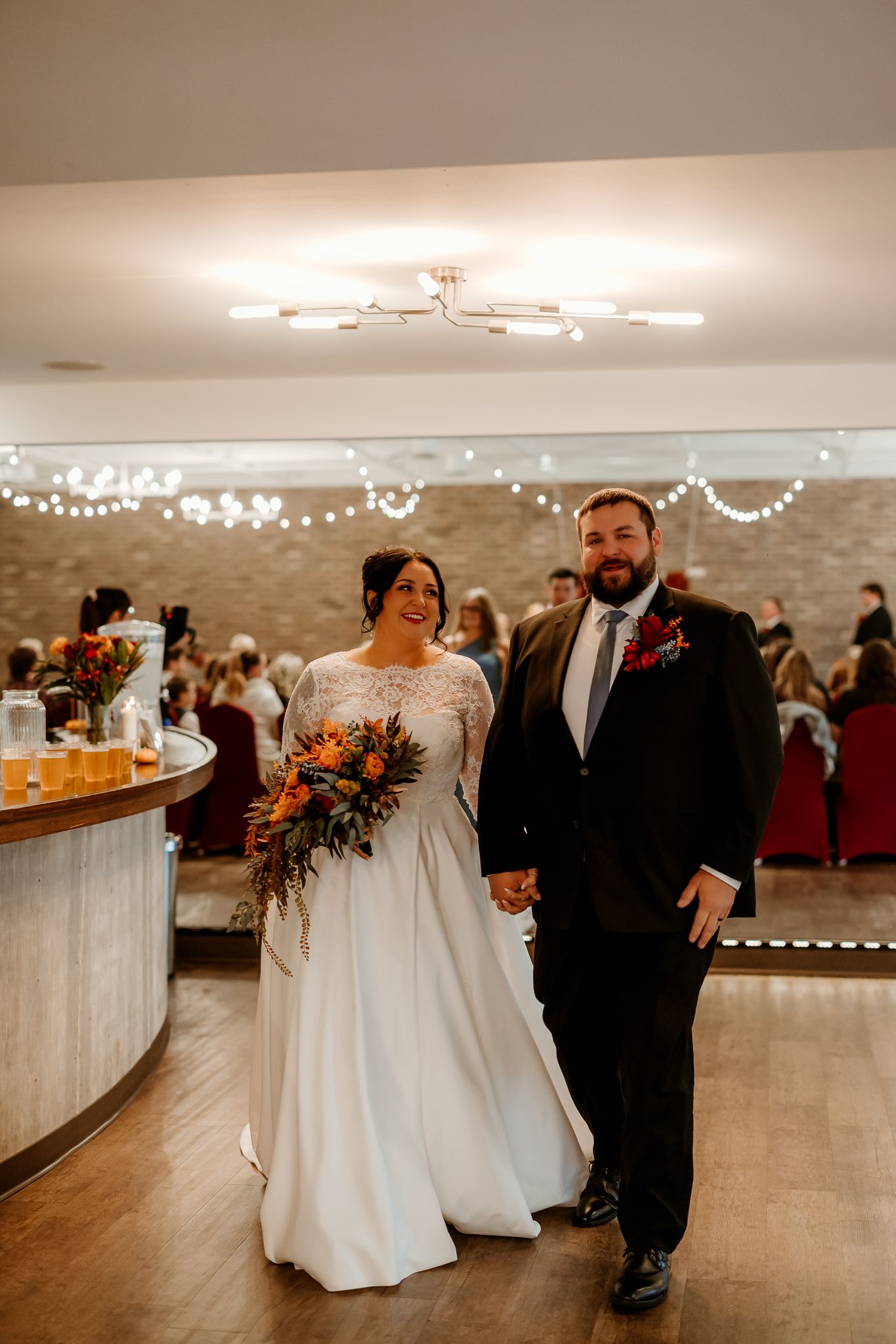 Smiling bride in a white lace gown and groom in a dark suit hold hands, walking through a wedding reception with an orange bouquet.