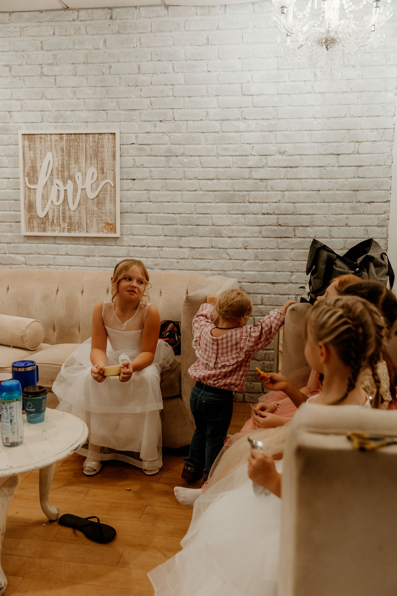 Girls in white dresses and a toddler gather on a tufted sofa in a bright room with a white brick wall and a framed love sign.