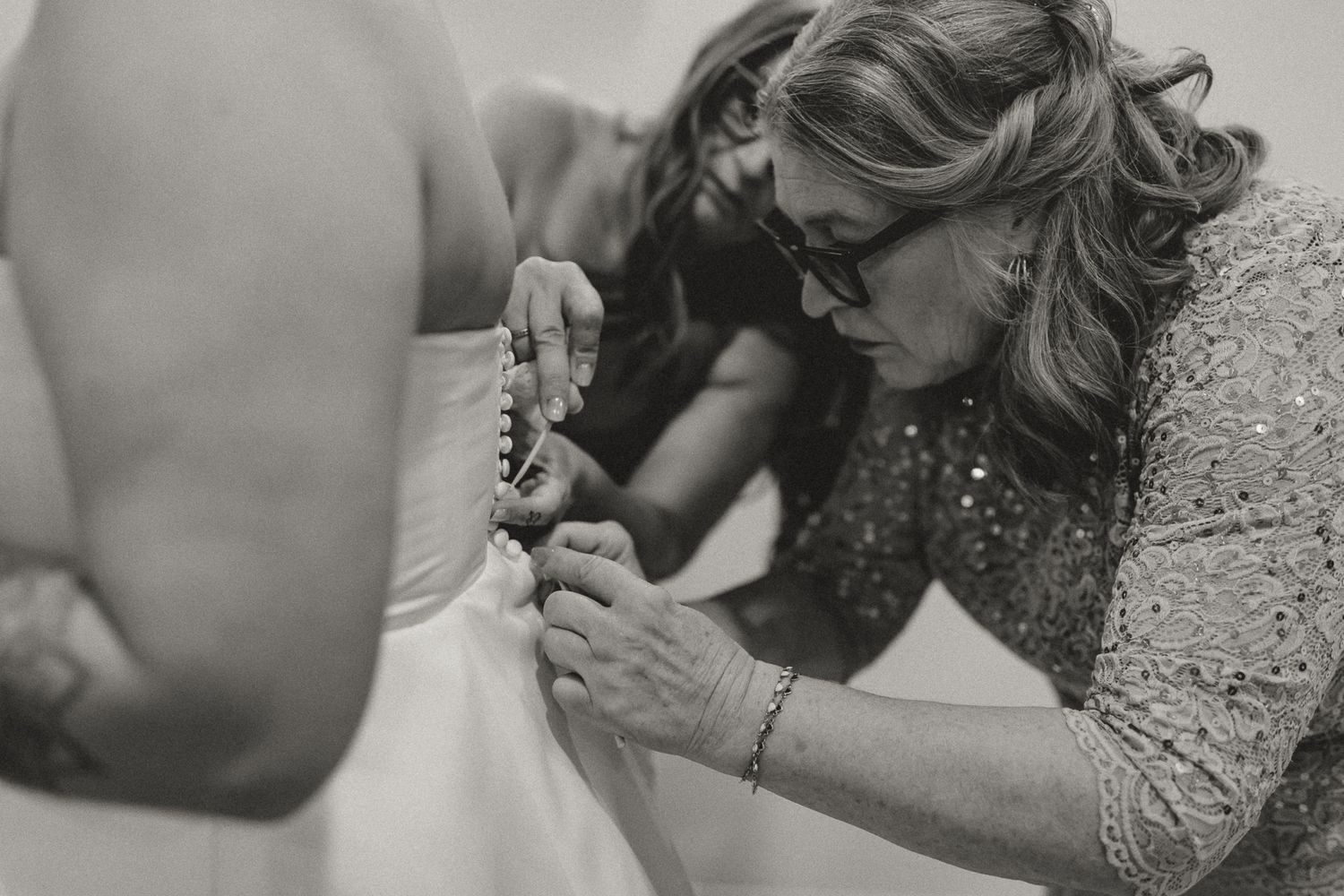 Woman in glasses and a sequined lace top helps a bride fasten the back of her wedding dress in a black and white photo.