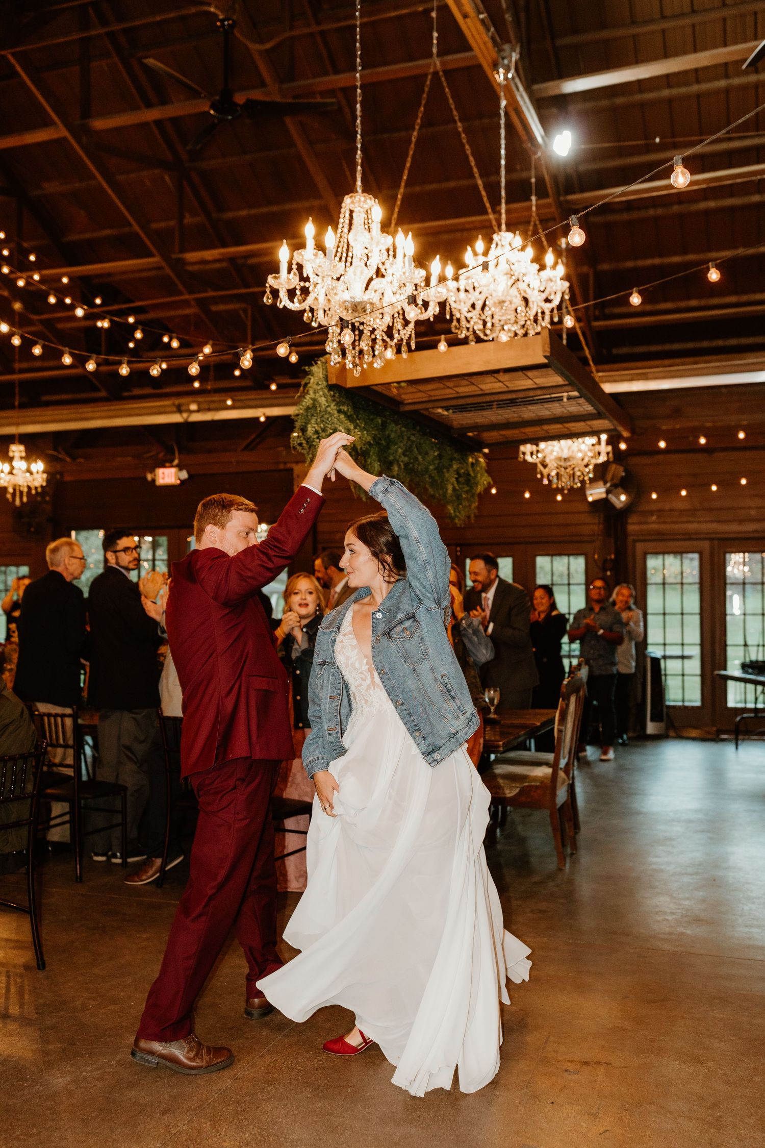 Couple dances at a rustic wedding reception, the bride in a denim jacket twirling beneath crystal chandeliers and string lights.
