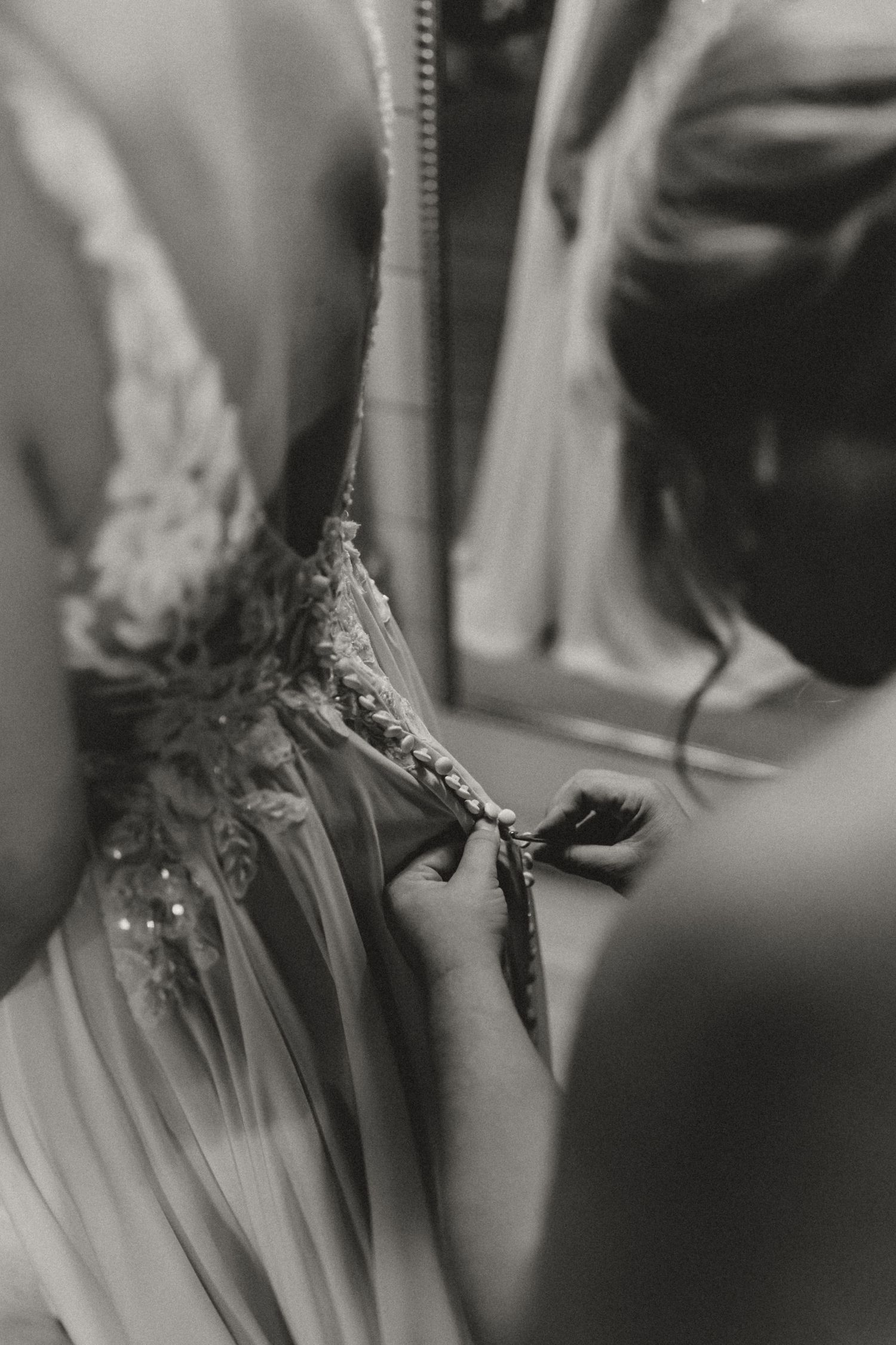 Black and white photo of a bridesmaid fastening the small buttons on the back of a lace wedding dress with beaded embellishments.