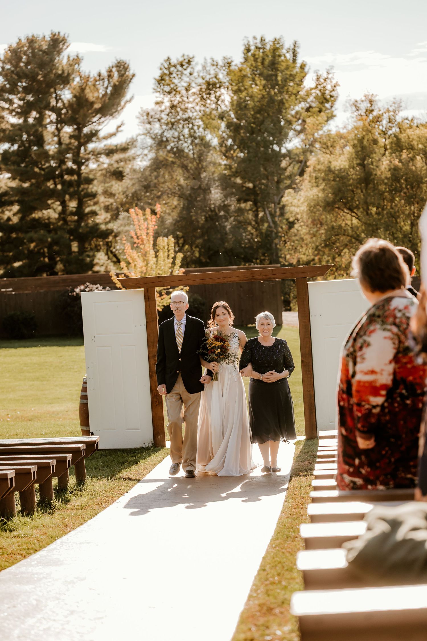 A bride in a flowing white gown walks down an outdoor wedding aisle escorted by an older man, with guests and trees in the background.
