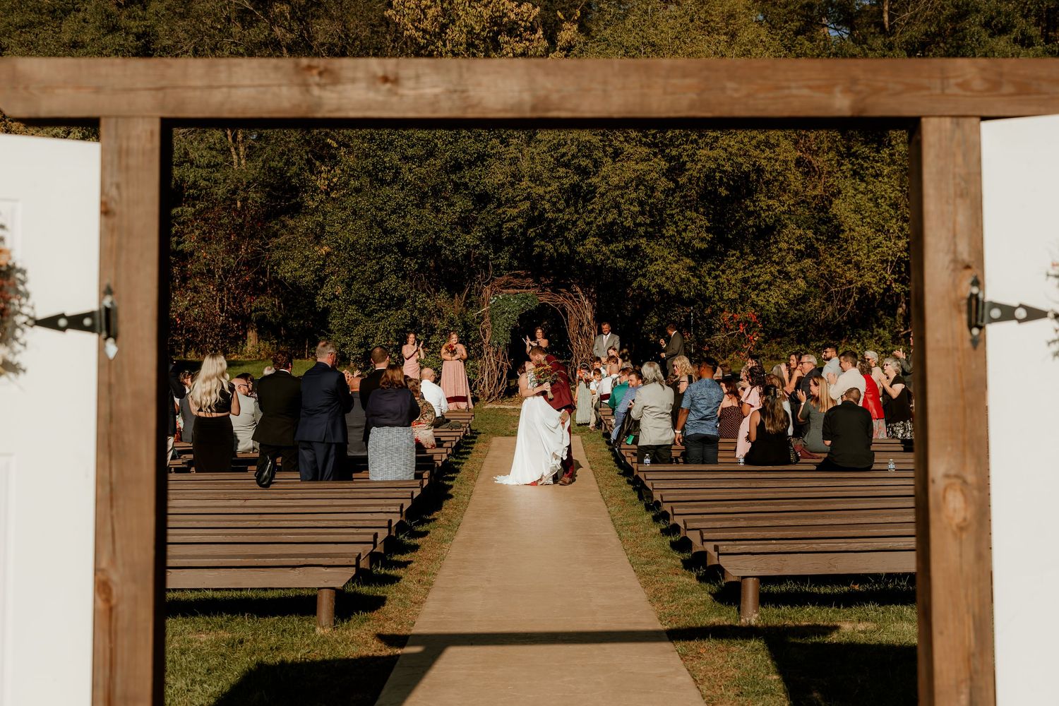 Bride in a white gown walks down the aisle of an outdoor wedding ceremony as guests watch from wooden benches framed by rustic barn doors.