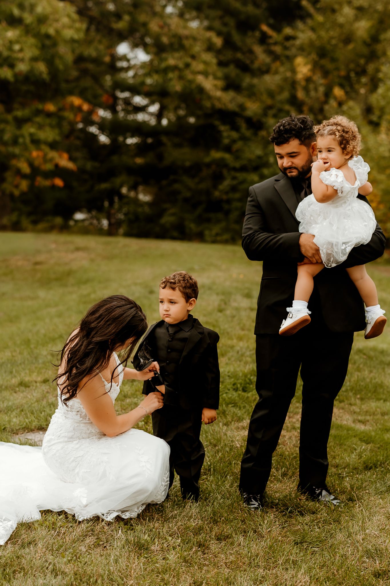 A bride in a white gown kneels on the grass adjusting a young boy's suit while a groom holds a toddler girl in a white dress.